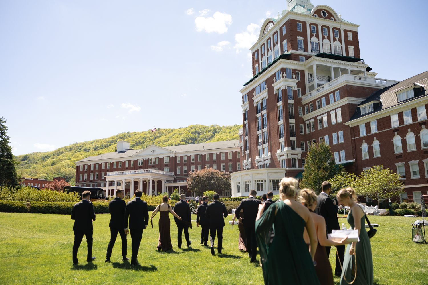 Wedding guests in formal attire gathered on lawn outside grand historic brick hotel building.