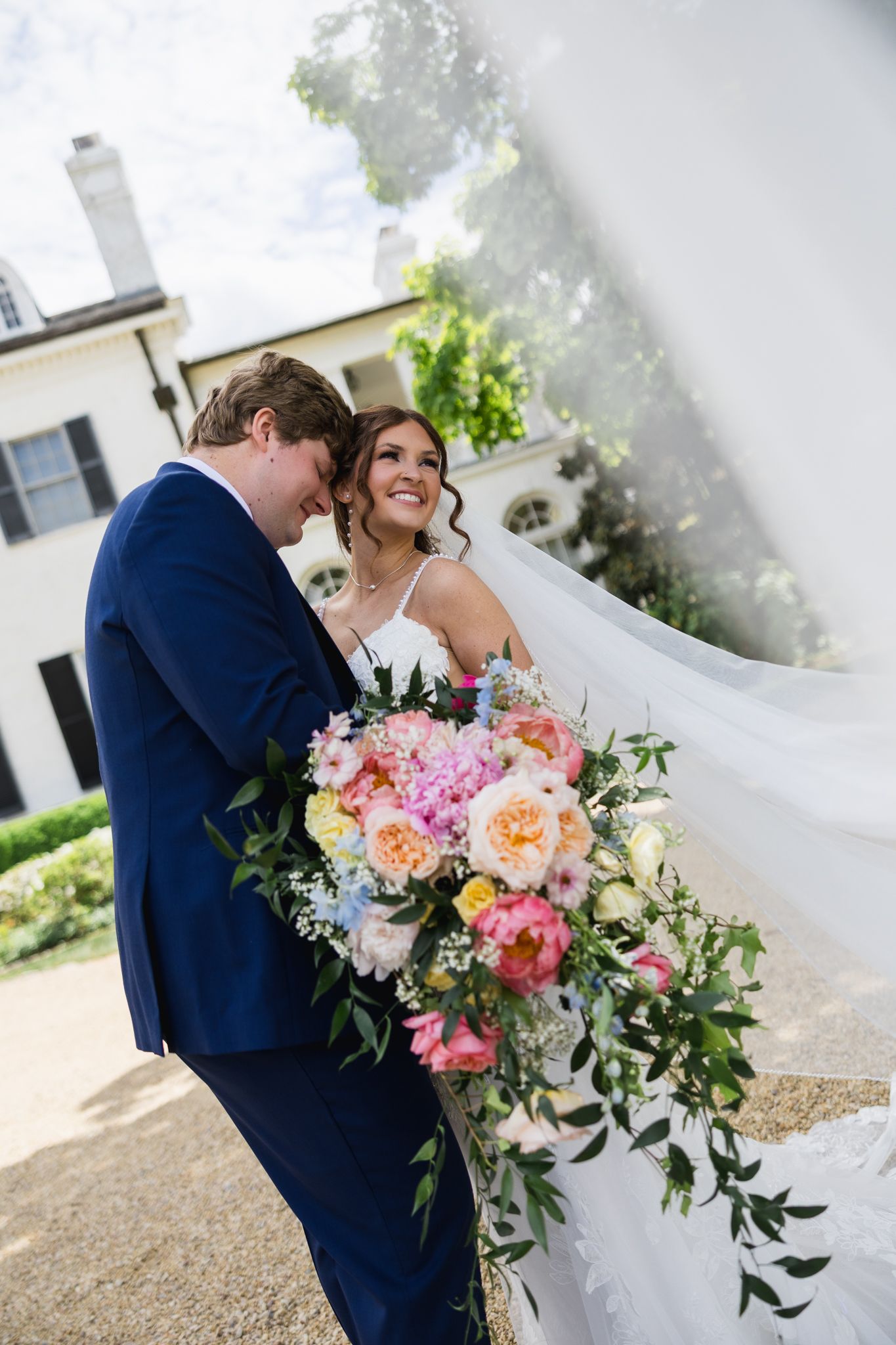 A smiling bride holding a colorful bouquet leans close to her groom in a navy suit outside a white manor.