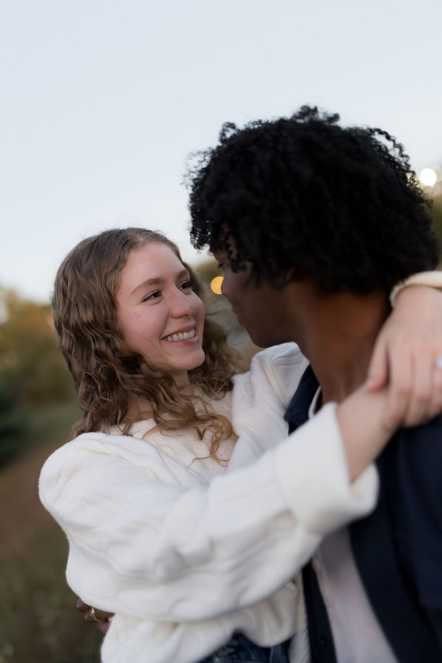 A smiling young woman embraces a curly-haired man outdoors at dusk in a romantic moment.