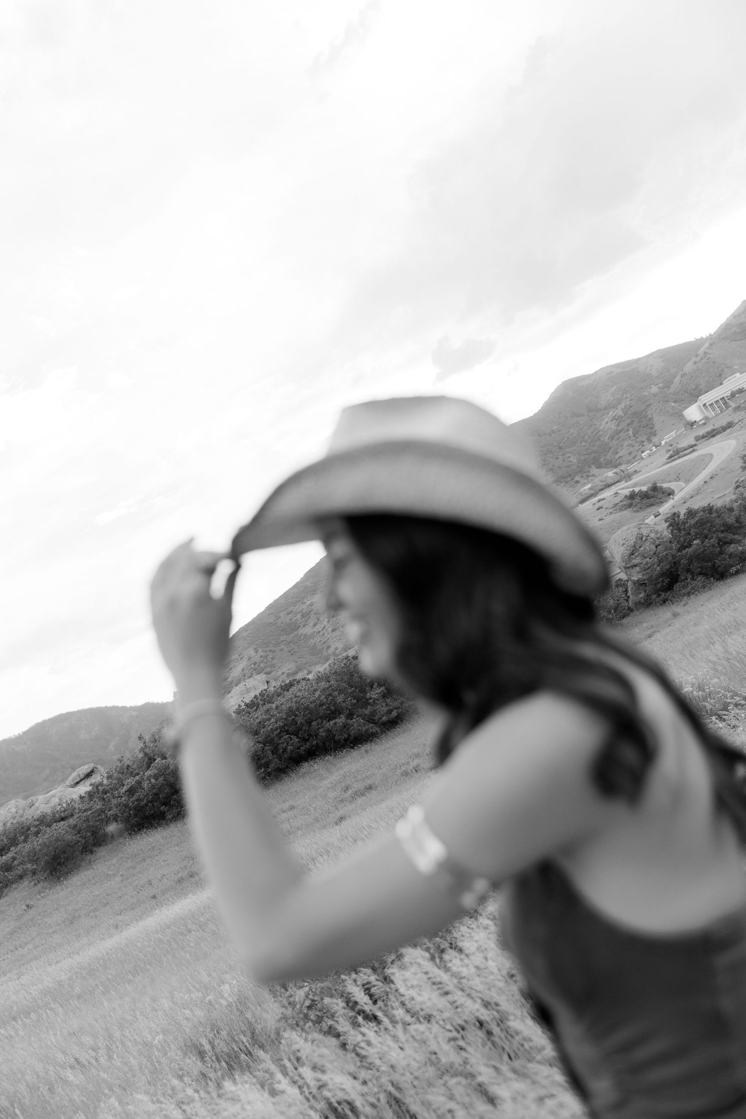 Black and white photo of a woman holding a cowboy hat in a rural mountain landscape field.