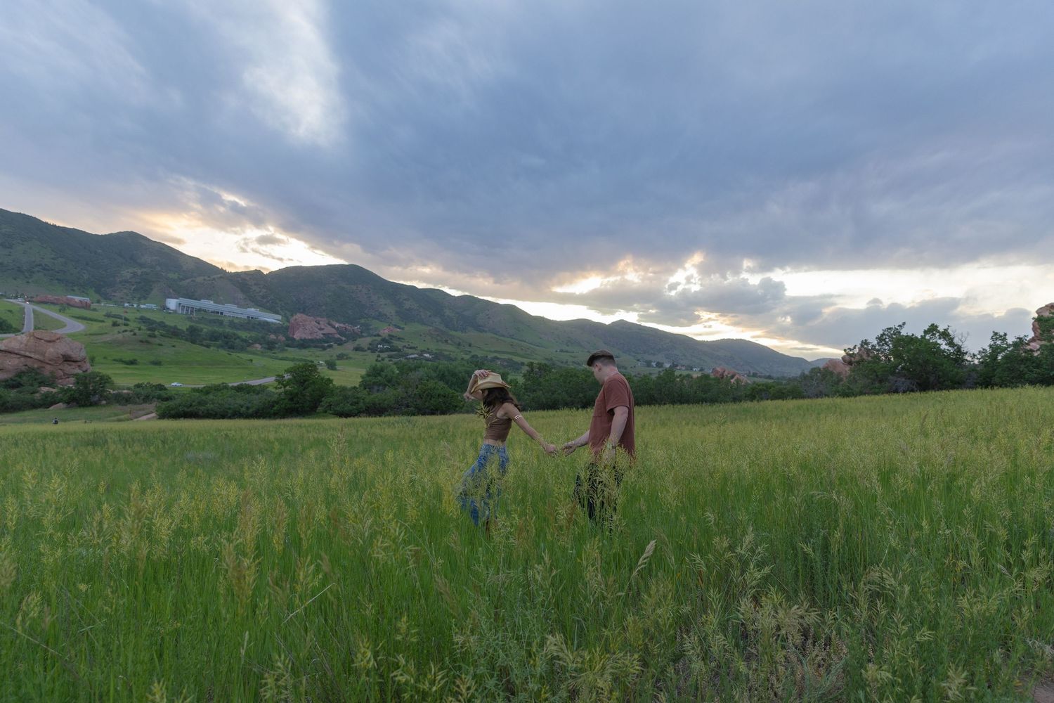 Couple walking hand in hand through tall green grass field with mountain sunset backdrop.