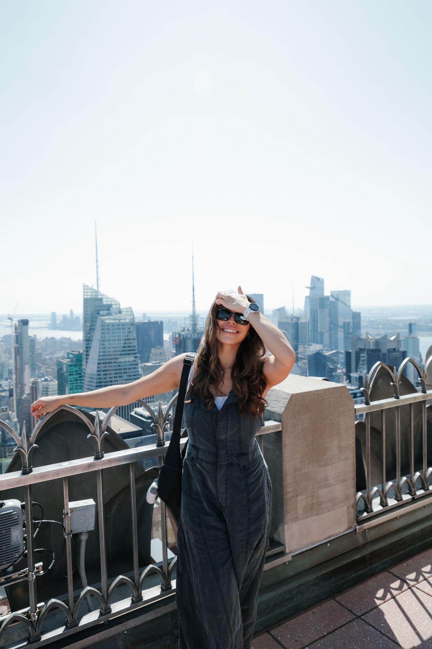 Smiling woman in denim jumpsuit and sunglasses posing on a rooftop overlooking a city skyline.