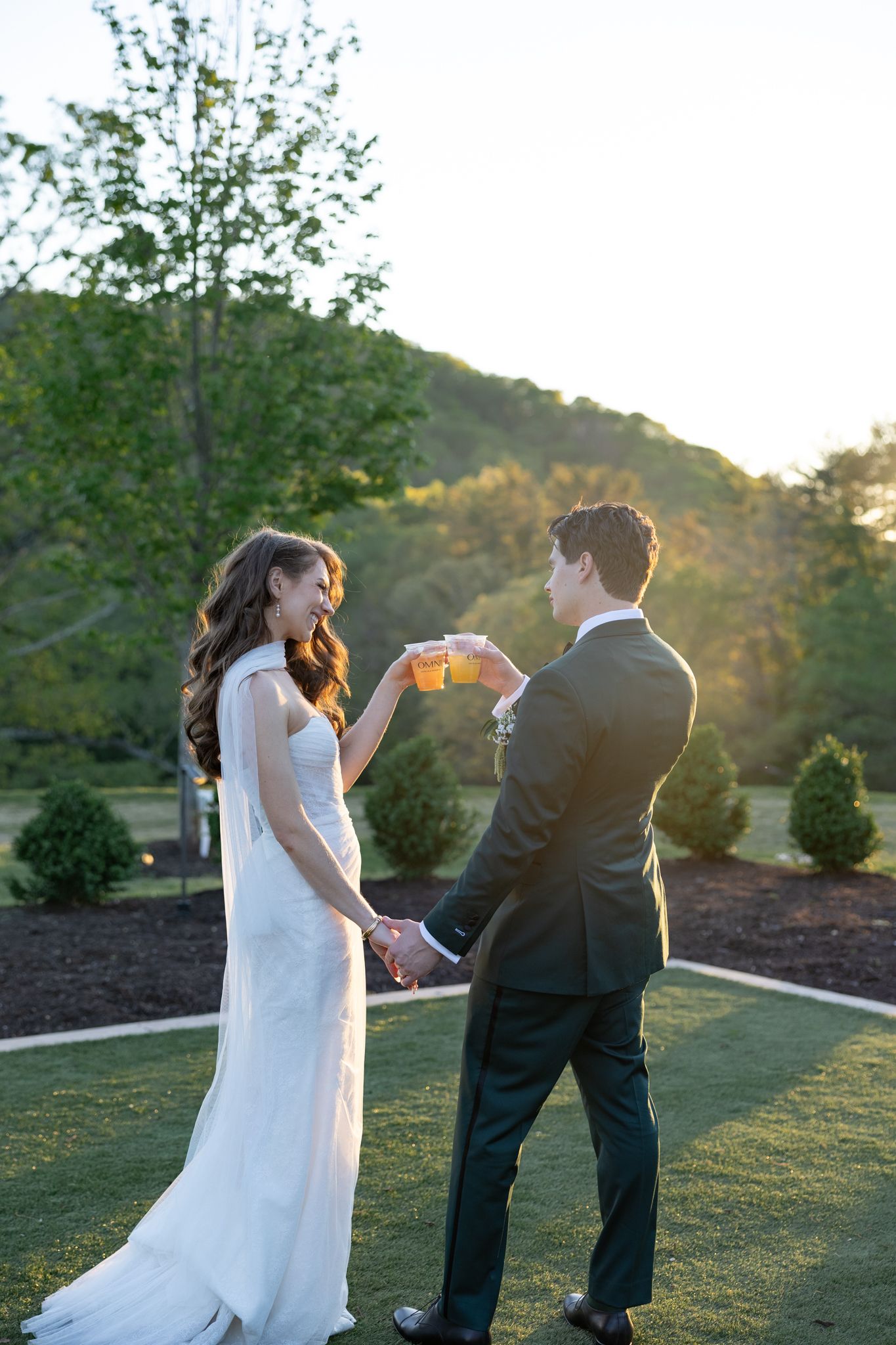 Bride and groom toasting with drinks outdoors at sunset, surrounded by lush greenery on their wedding day.