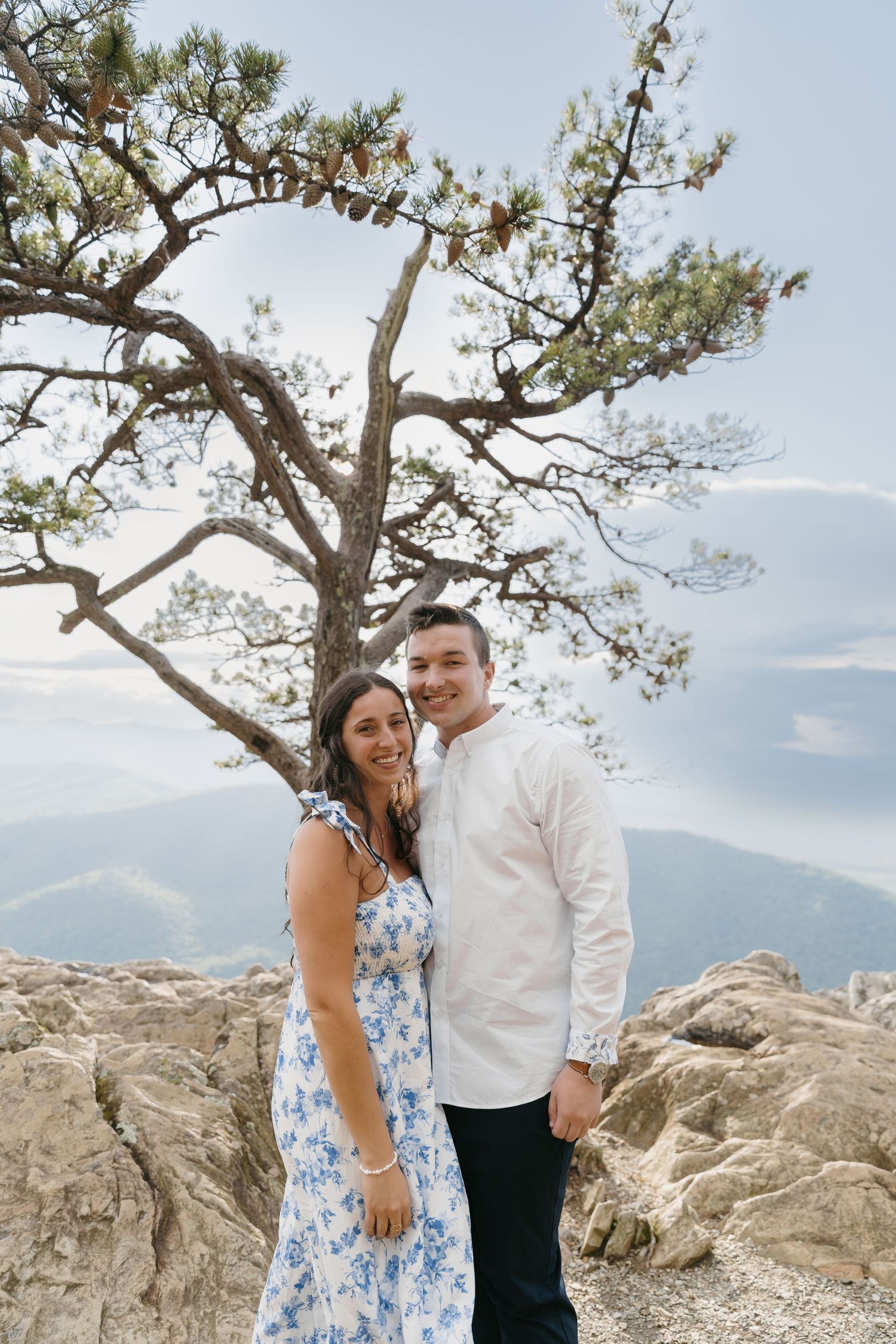 Smiling couple embracing on mountain rocks under a pine tree with misty hills behind.