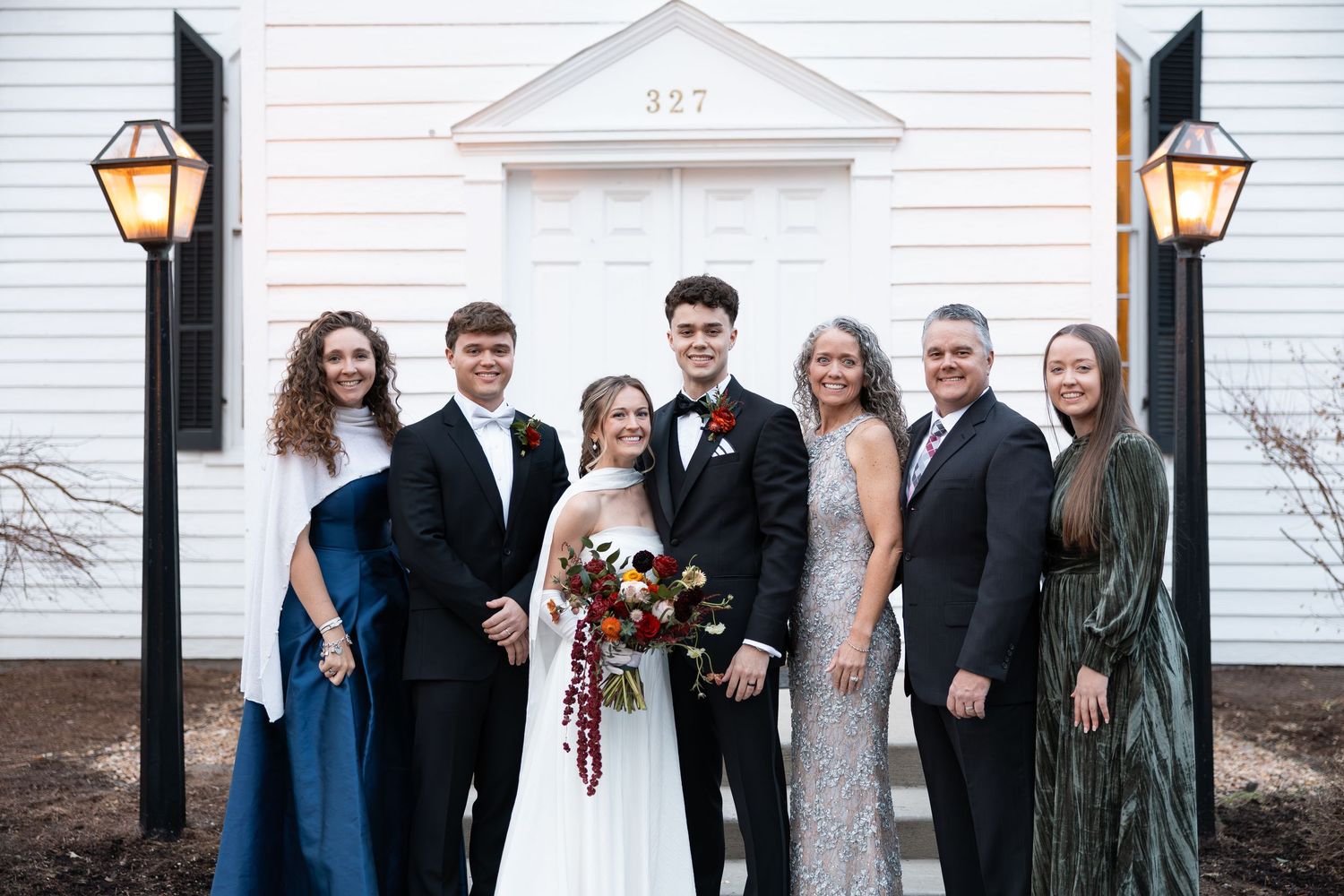 Wedding party of seven posing together in front of a white chapel with black shutters.