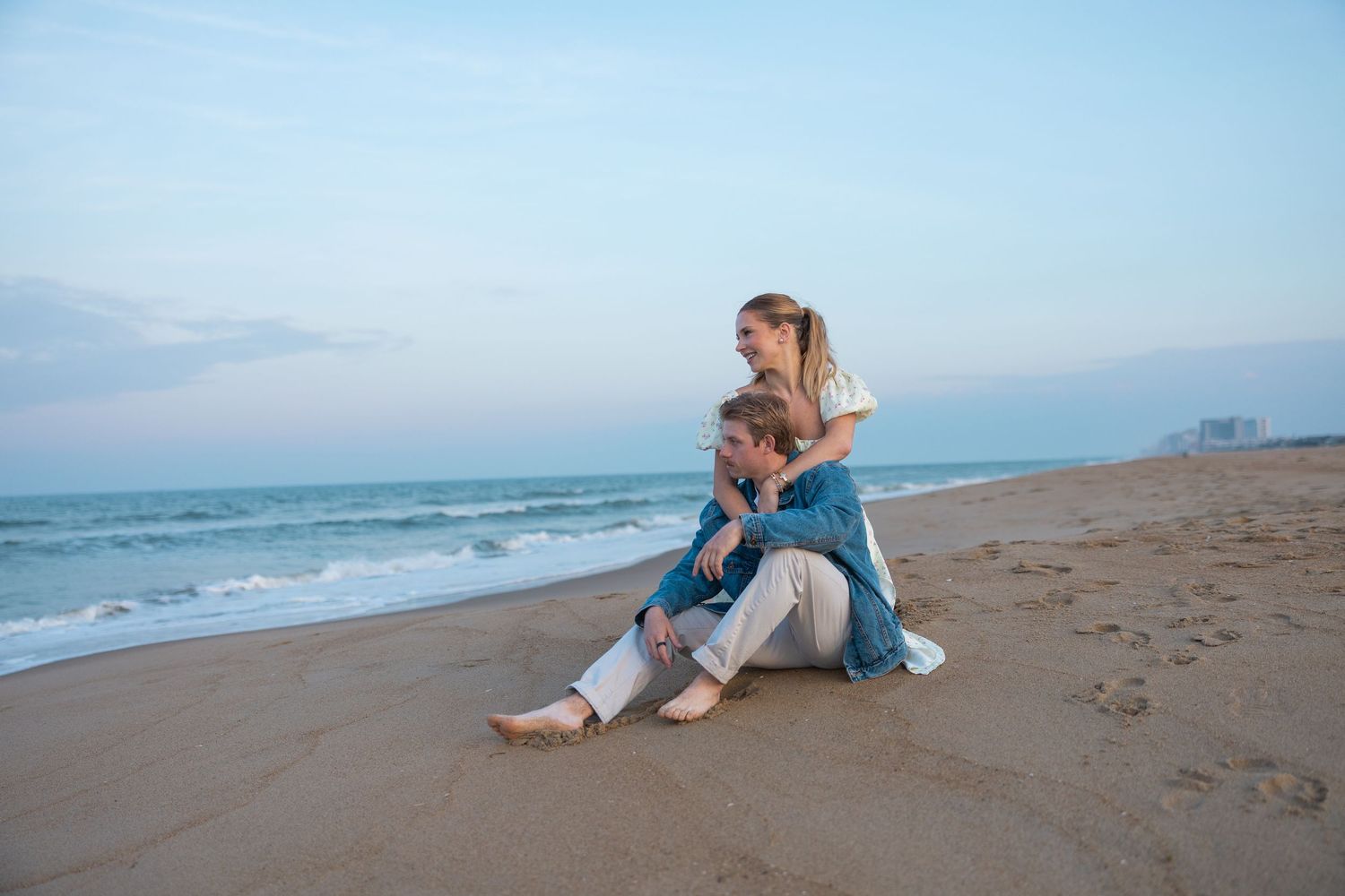 Couple sitting together on sandy beach at dusk, woman embracing man from behind with ocean and city skyline behind them.
