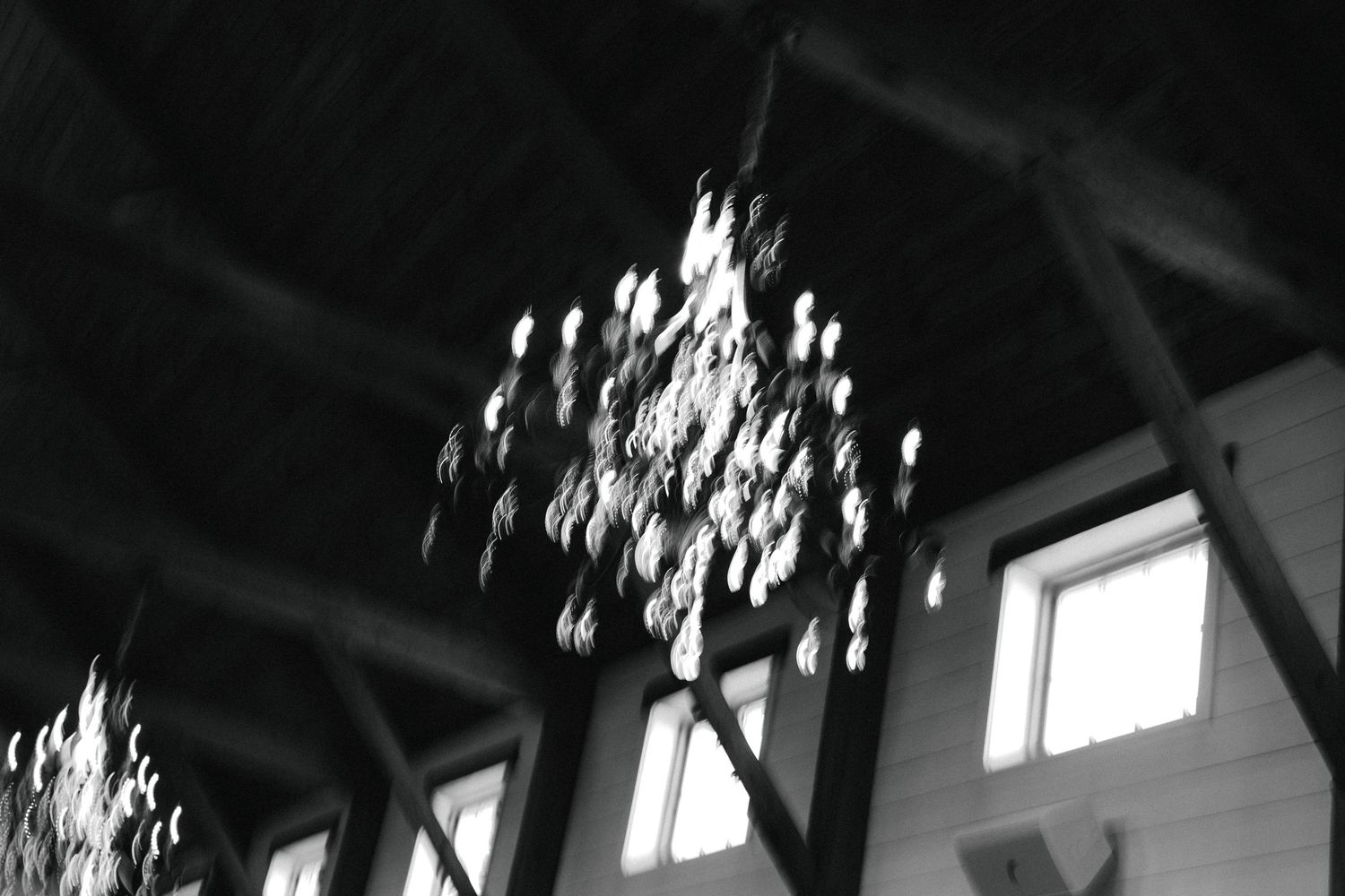 Black and white photo of an elegant chandelier hanging from rustic wooden ceiling beams in a wedding venue.