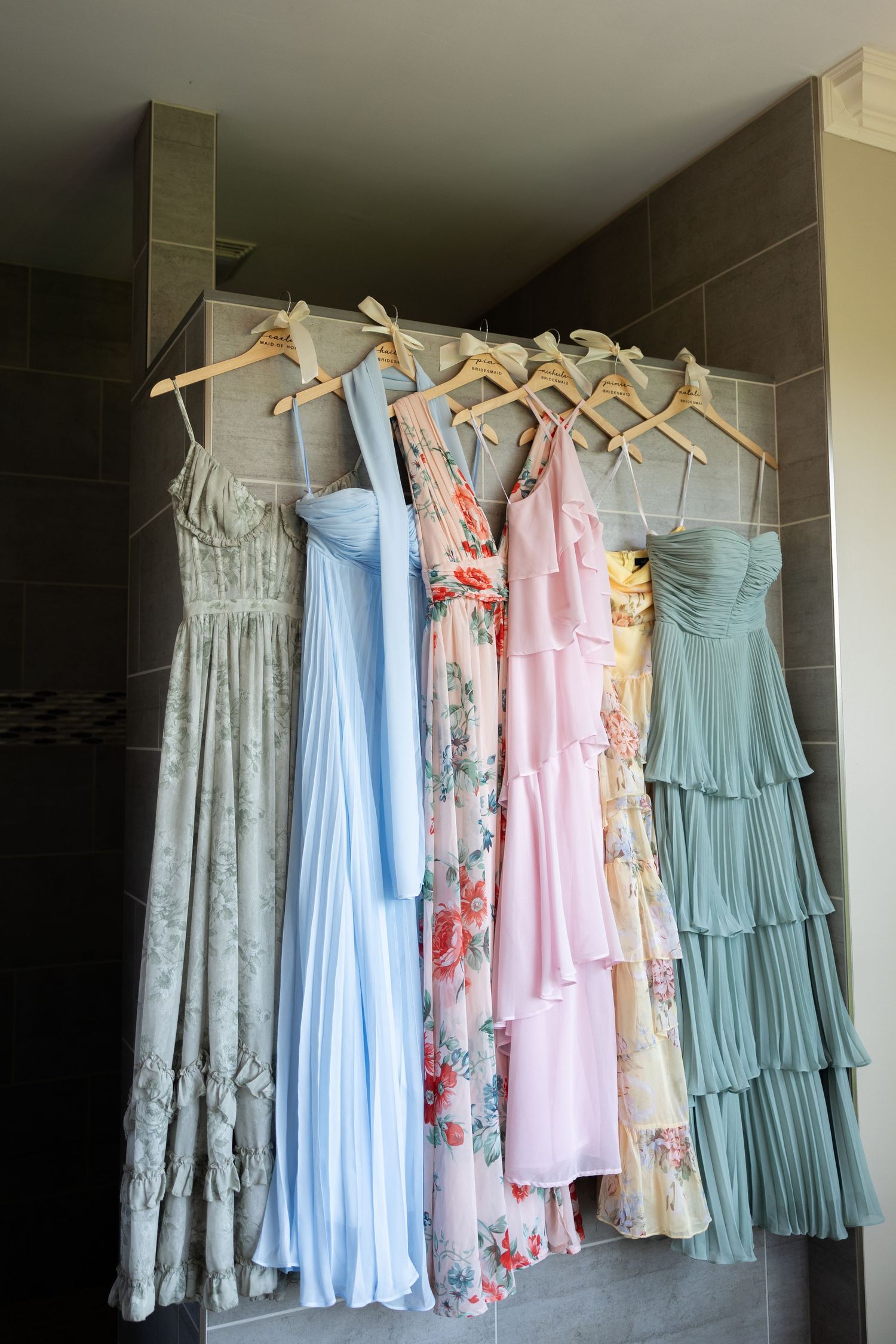 Six pastel bridesmaid dresses on wooden hangers displayed against a tiled wall in soft pink, blue, and floral patterns.