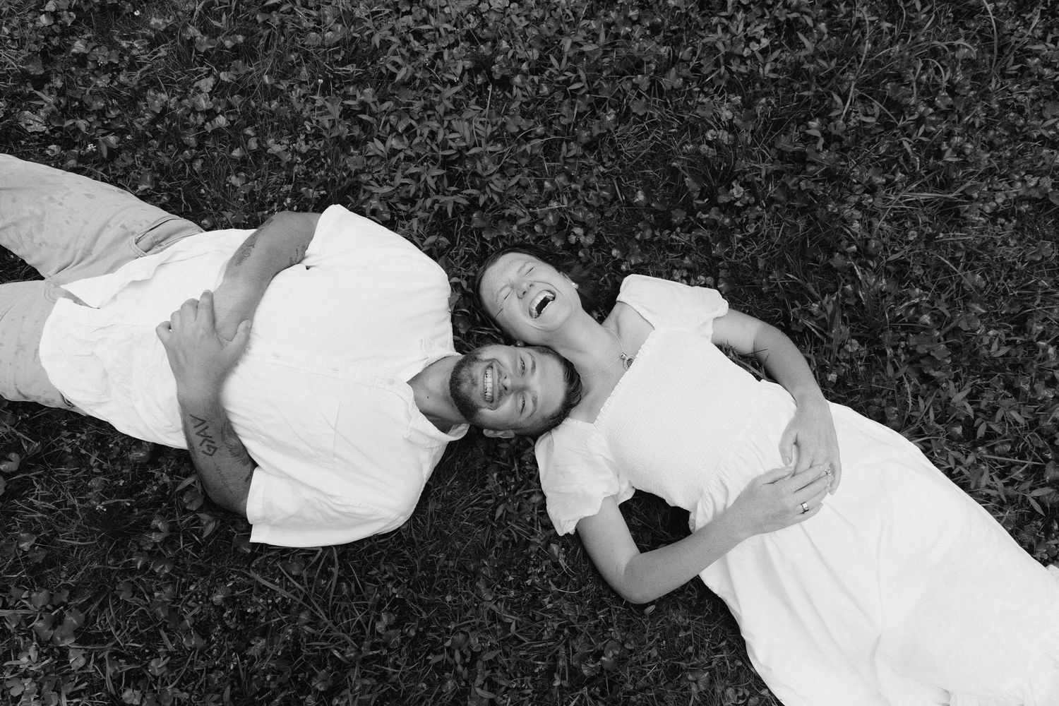 Black and white photo of a couple lying on grass, heads together, smiling and embracing outdoors.