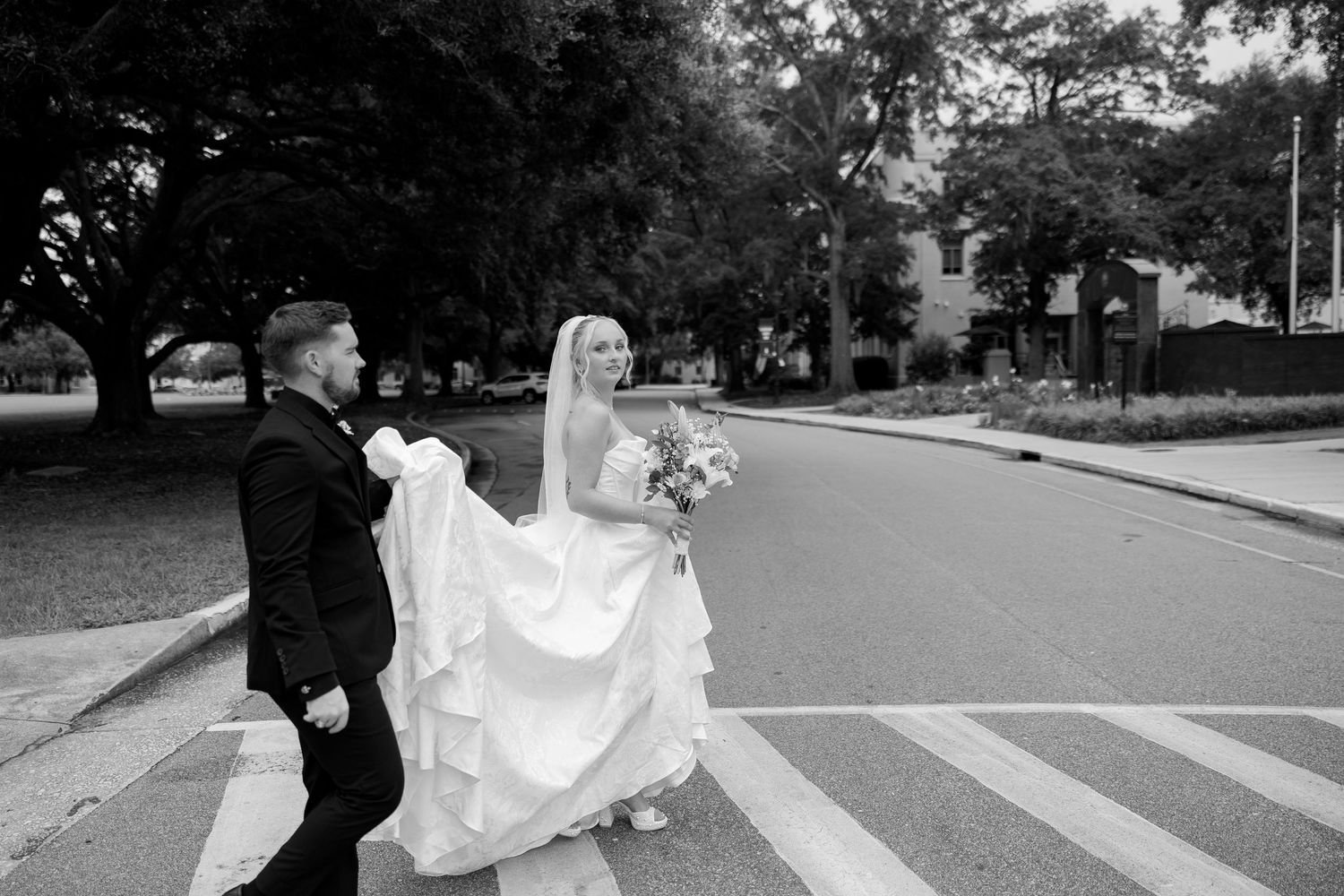 Black and white photo of a bride and groom crossing a street, groom holding the bride's dress train.