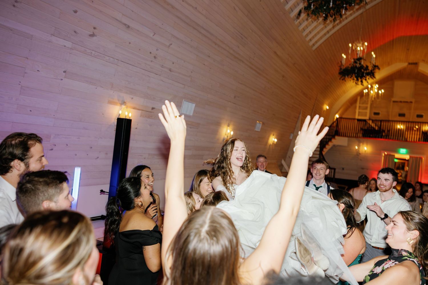 Bride in white gown is lifted by wedding guests during lively reception dance floor celebration.