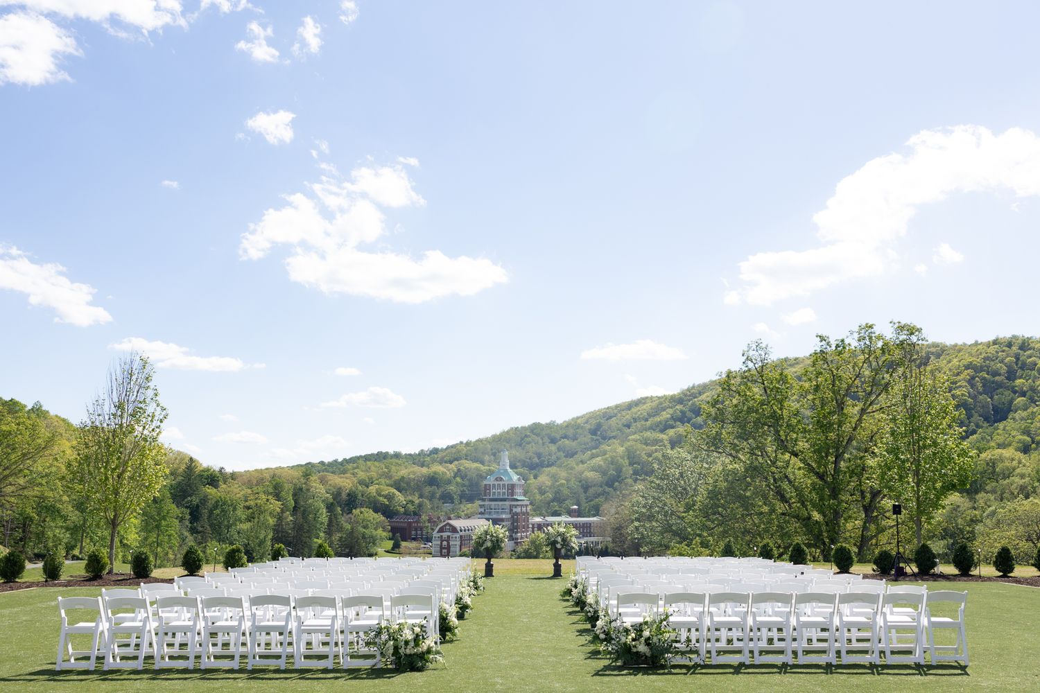 Outdoor wedding ceremony setup with white chairs on green lawn, surrounded by trees and hills under a clear blue sky.