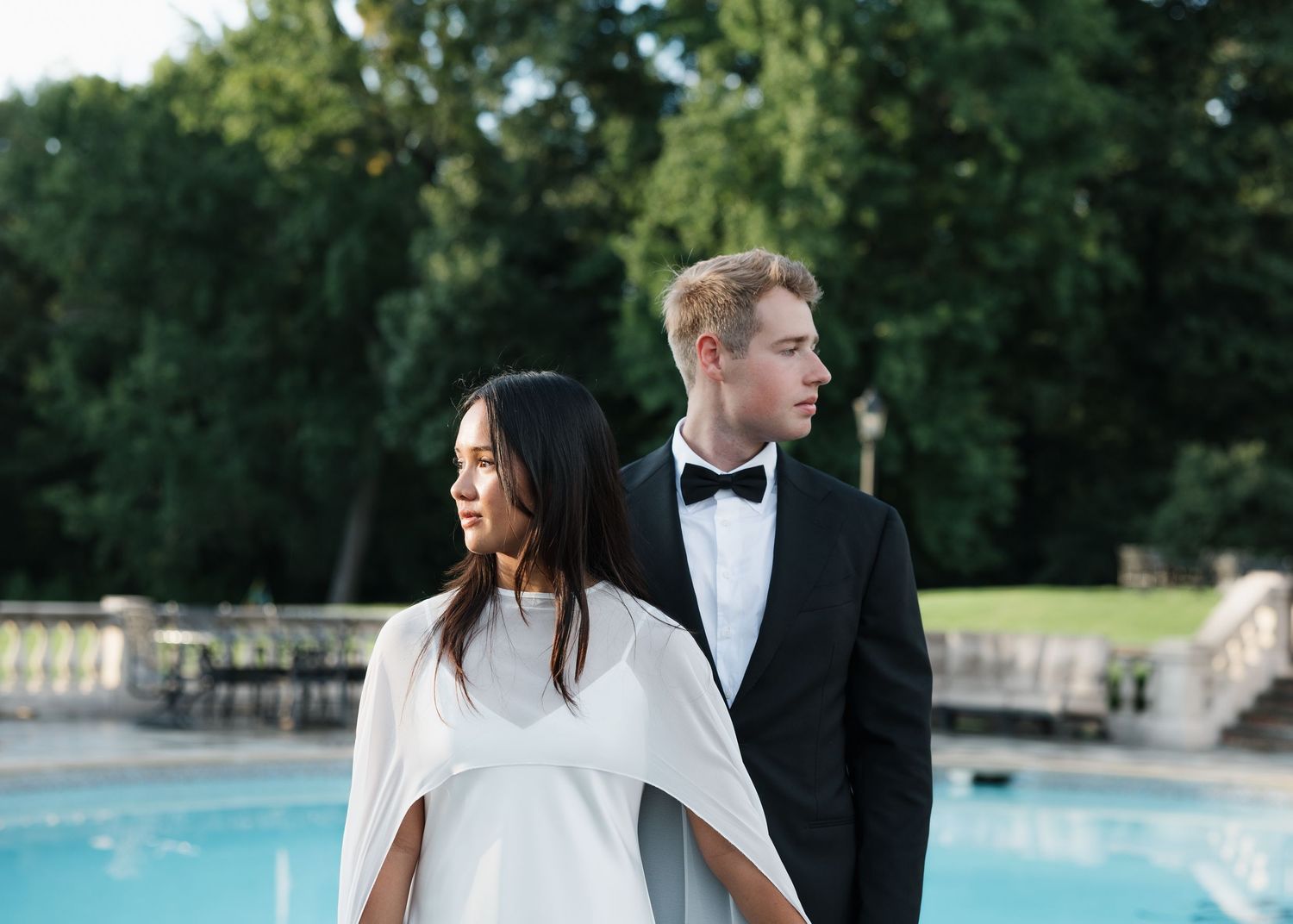 Elegant couple in formal attire standing by a pool, woman in white cape dress and man in black tuxedo with bow tie.