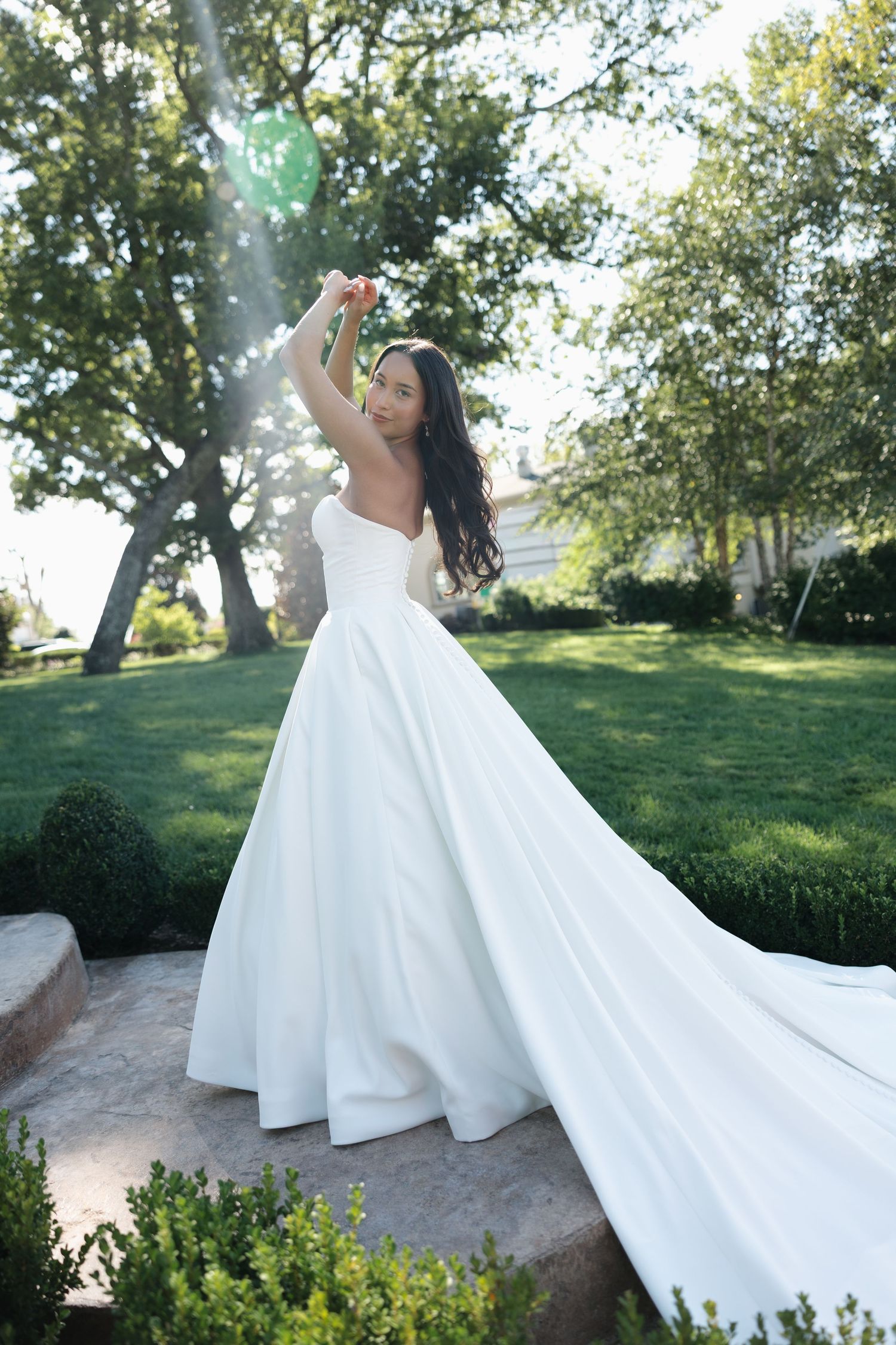 Bride in a flowing white ball gown with a long train poses gracefully outdoors in a sunlit garden.