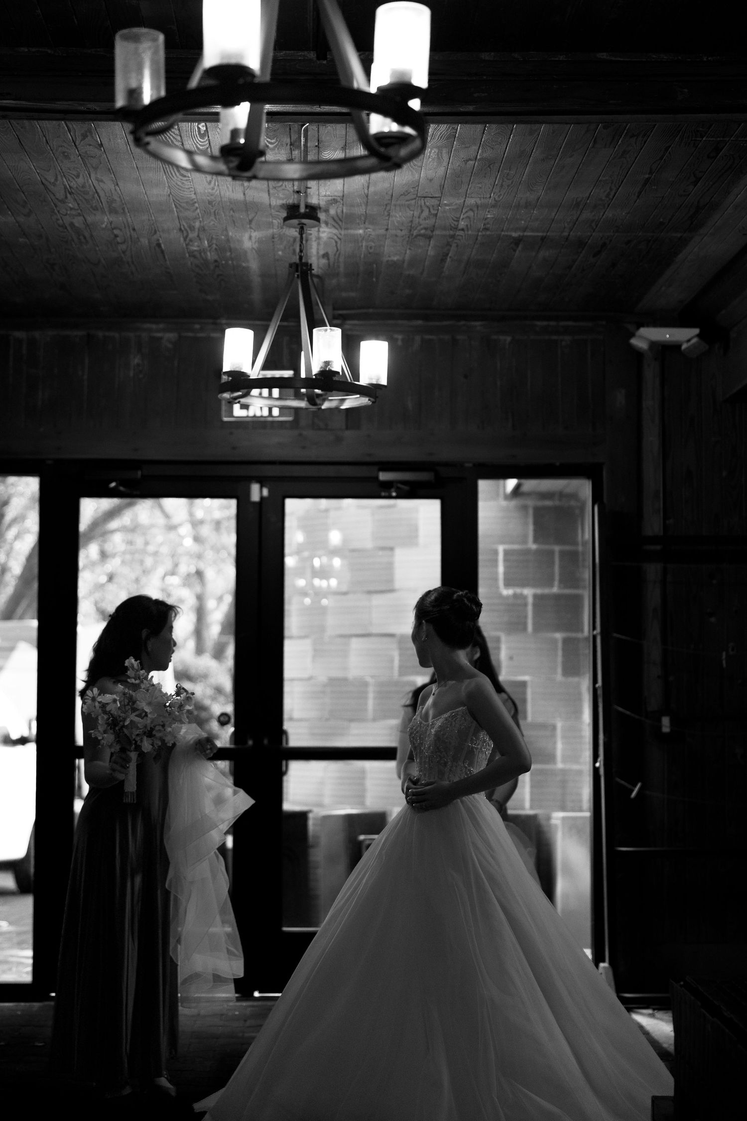 Black and white photo of a bride in a ballgown with bridesmaid holding flowers near elegant chandelier.