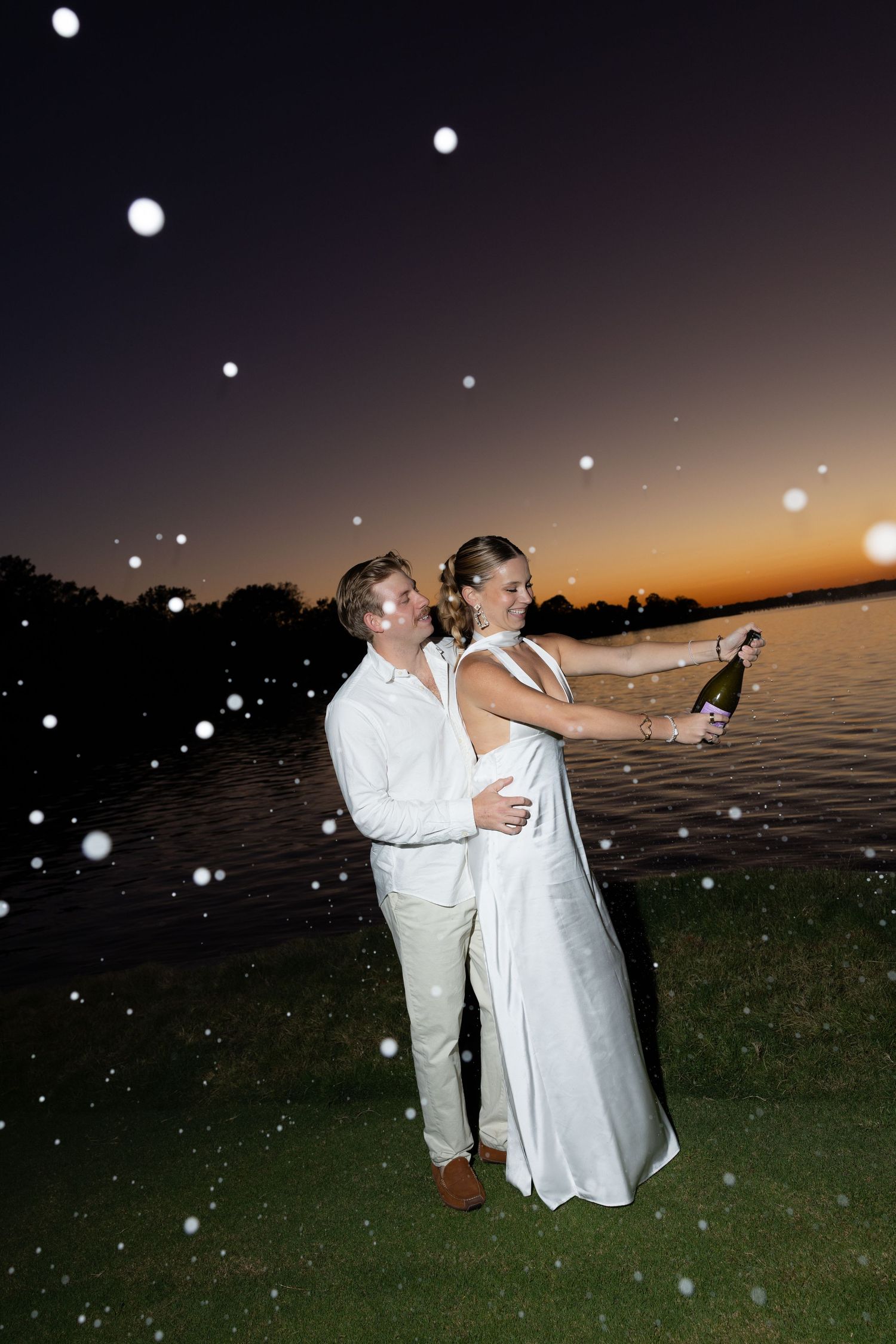Couple in white celebrating at sunset by the water, popping champagne with sparkling bokeh lights around them.