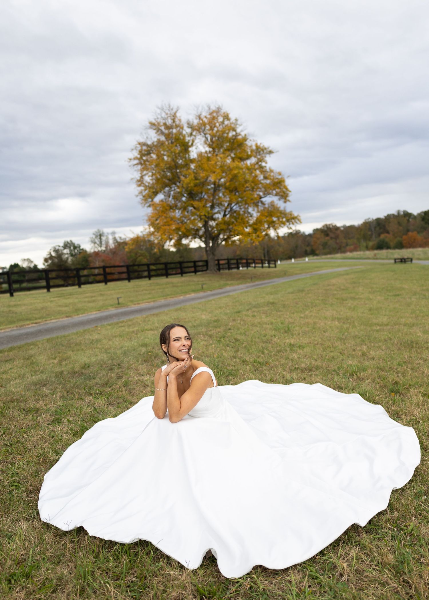 Bride in a flowing white ball gown sits gracefully in an open field with a golden autumn tree backdrop.