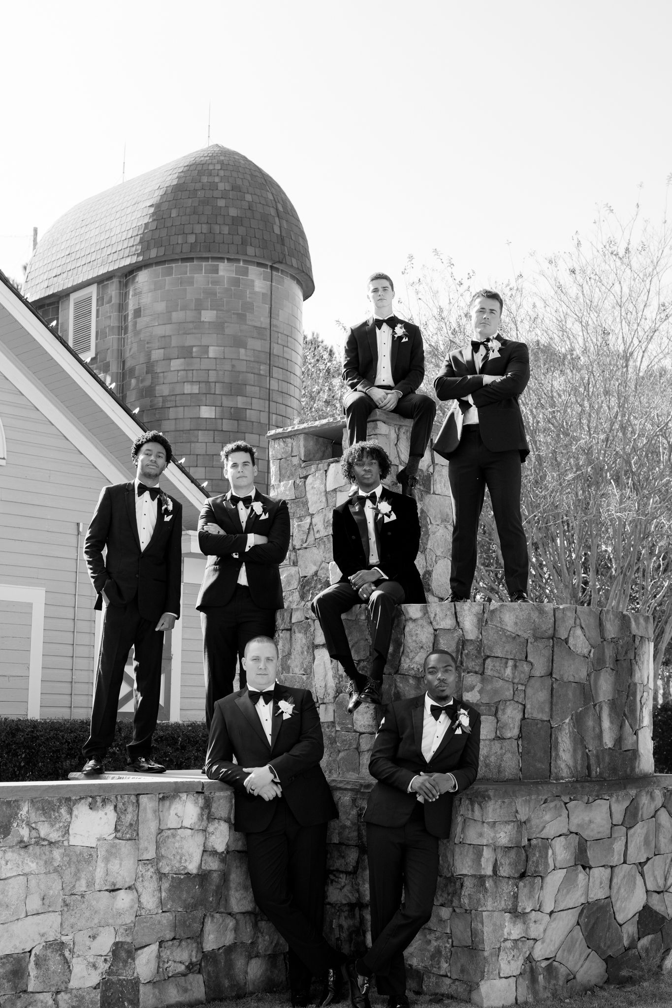 Groomsmen in black tuxedos posing on stone wall with rustic barn silo backdrop in black and white.