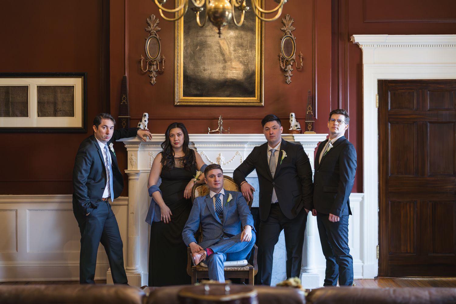Groomsmen and bridesmaids posing dramatically in elegant wood-paneled room with chandelier and fireplace.