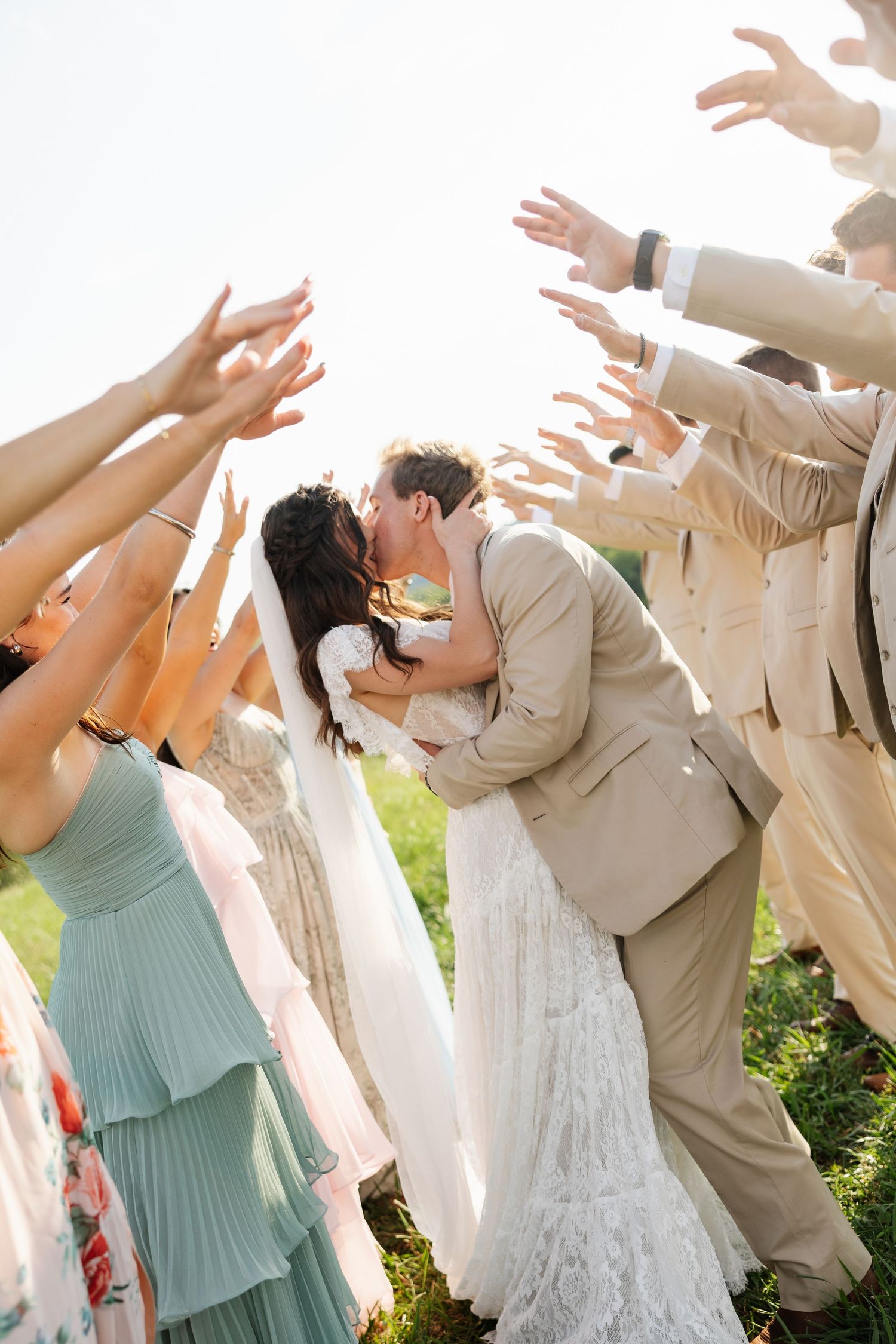 Bride and groom share a romantic dip kiss surrounded by cheering wedding guests raising their hands in celebration outdoors.