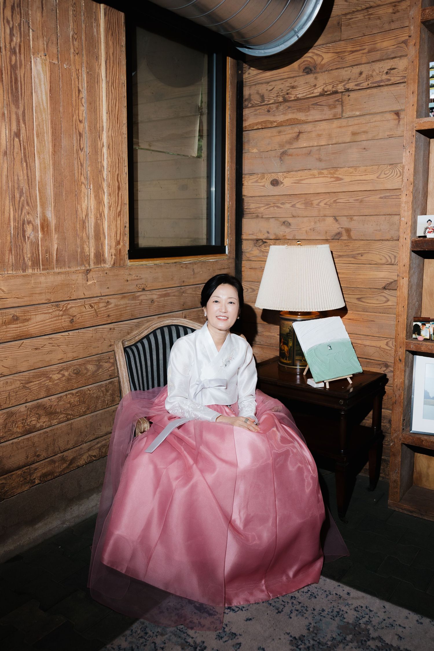 Woman in traditional Korean hanbok with pink skirt sits smiling in a wooden interior beside a lamp and desk.
