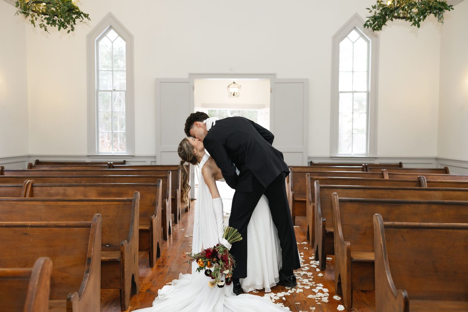Bride and groom share a romantic dip kiss in a white chapel aisle lined with wooden pews and rose petals.