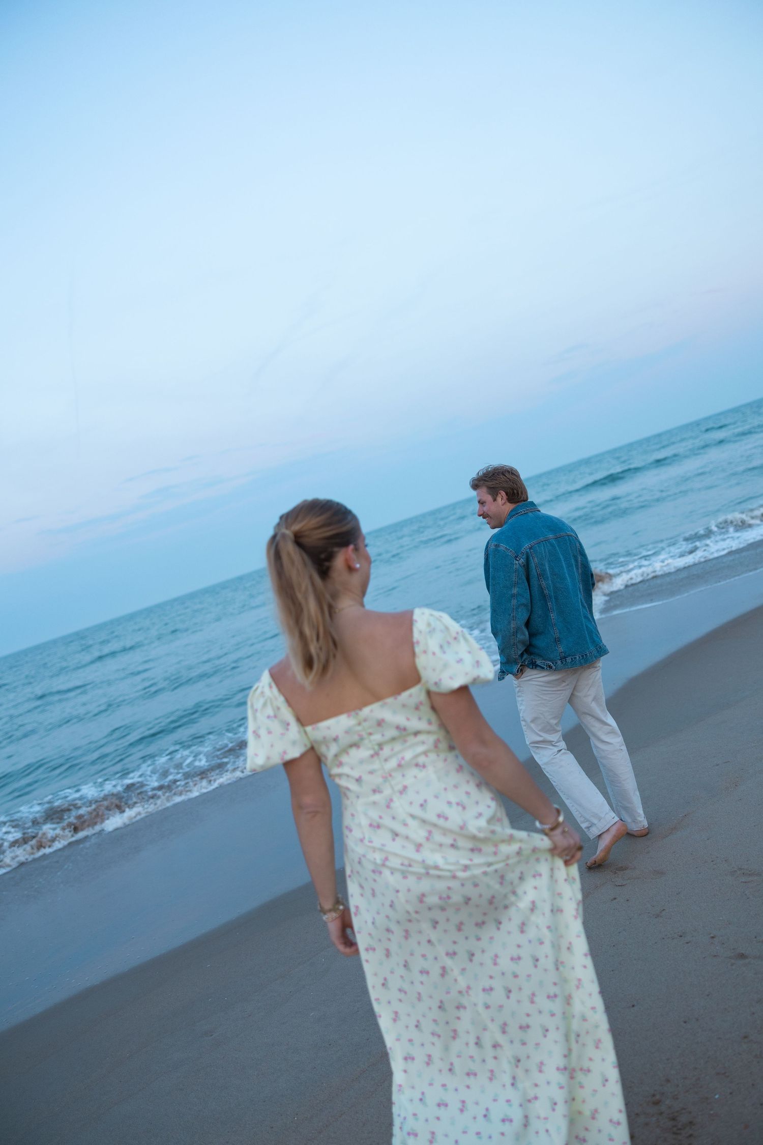 Woman in floral dress walks toward man in denim jacket on a serene beach at dusk.