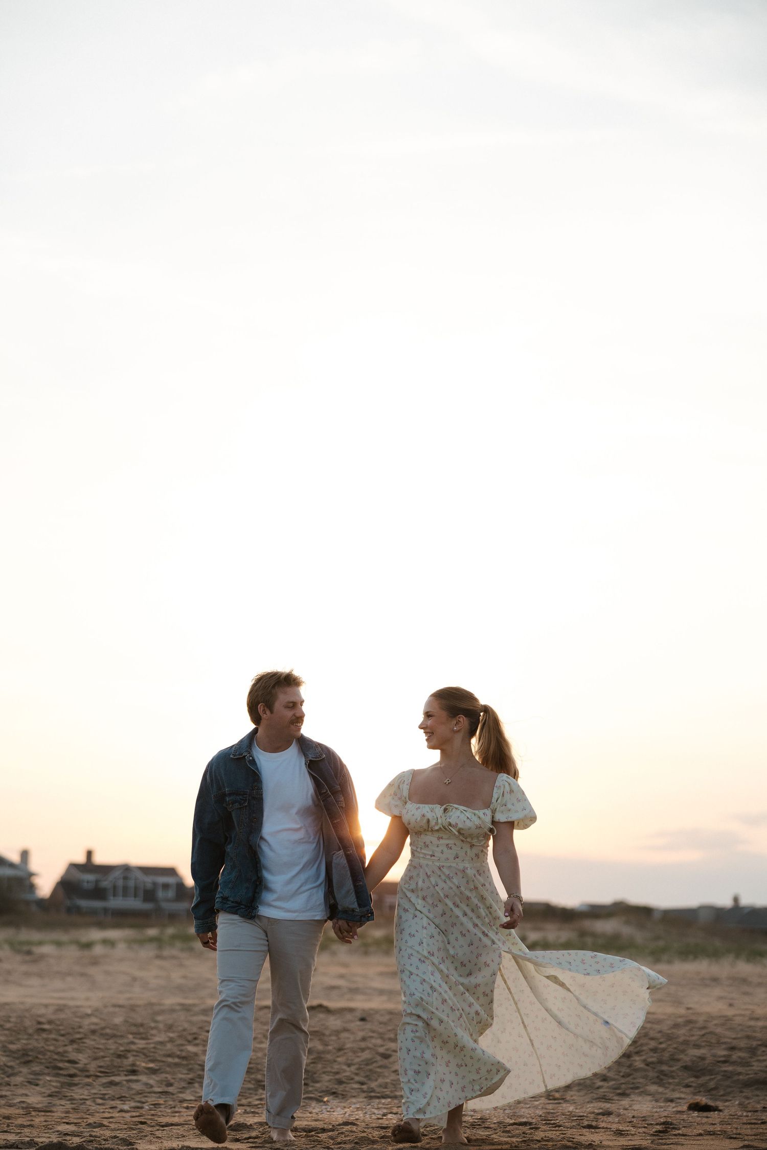 Couple holding hands and walking on sandy beach at sunset, woman in flowing floral dress and man in denim jacket.