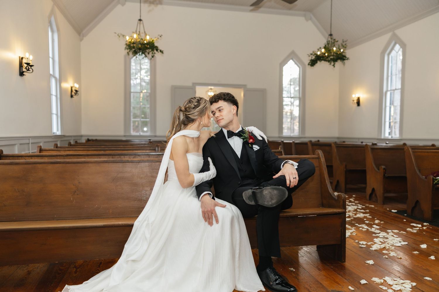 A bride and groom sit closely together on a church pew, sharing a romantic moment.