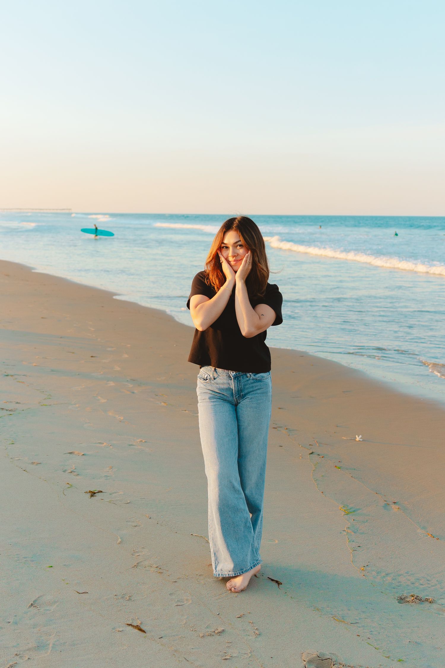 Woman wearing glasses in black crop top and blue wide-leg jeans poses on beach at golden hour with ocean backdrop.