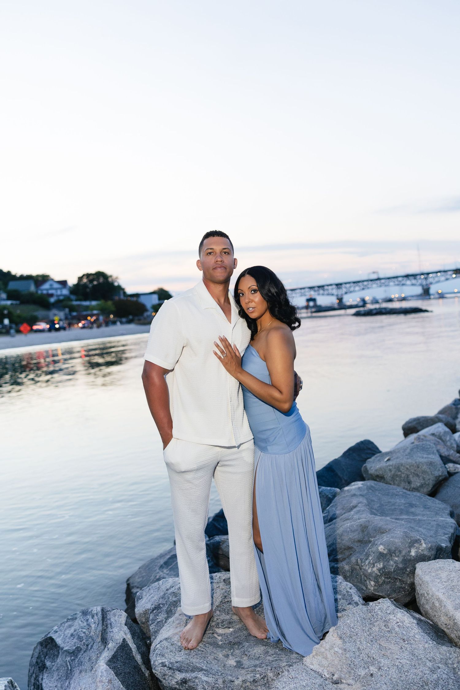Couple posing on rocky shoreline at dusk, woman in blue gown embracing man in white outfit near calm water.