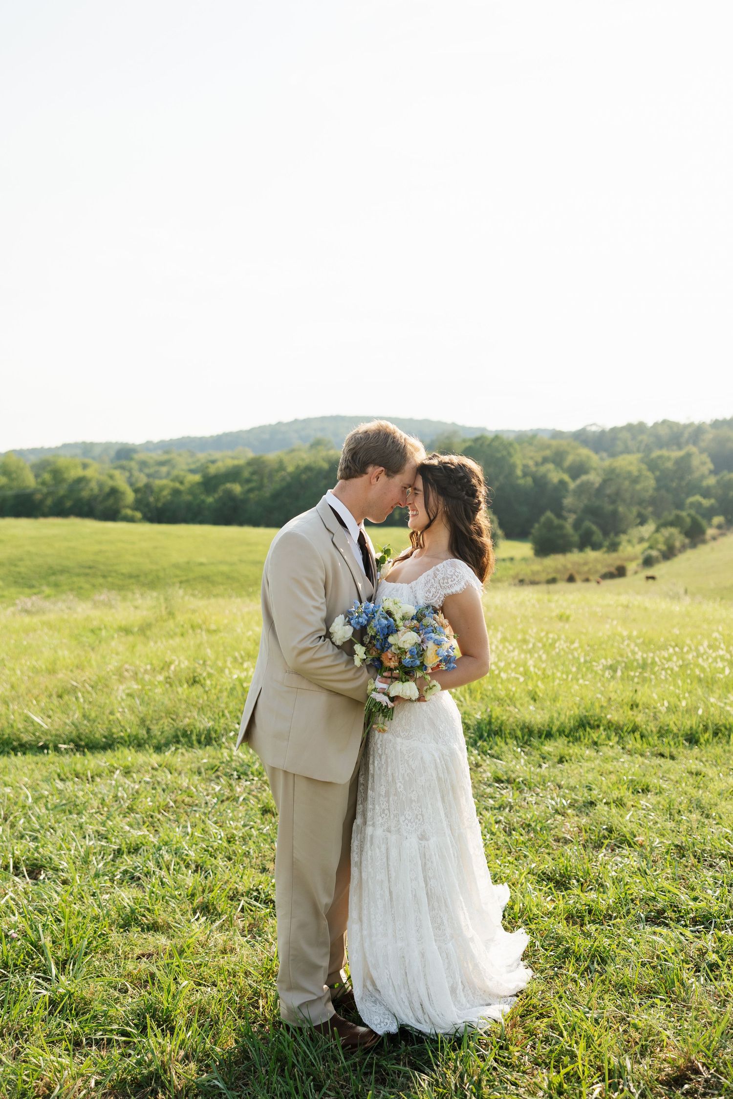 Bride and groom share a romantic moment in a lush green field, bride holding blue and white bouquet.