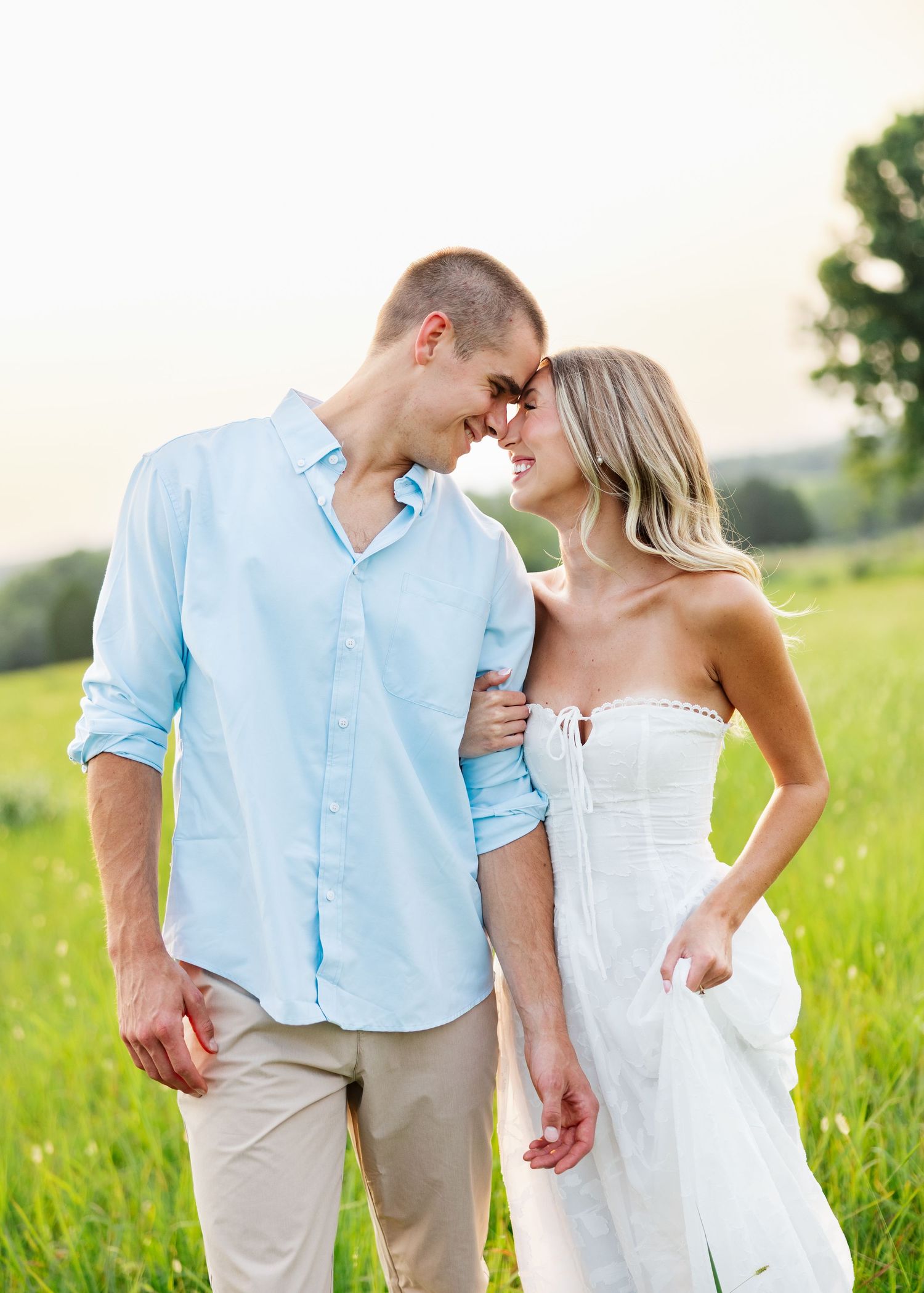 Romantic couple in a green field, man in light blue shirt and woman in white dress touching foreheads.