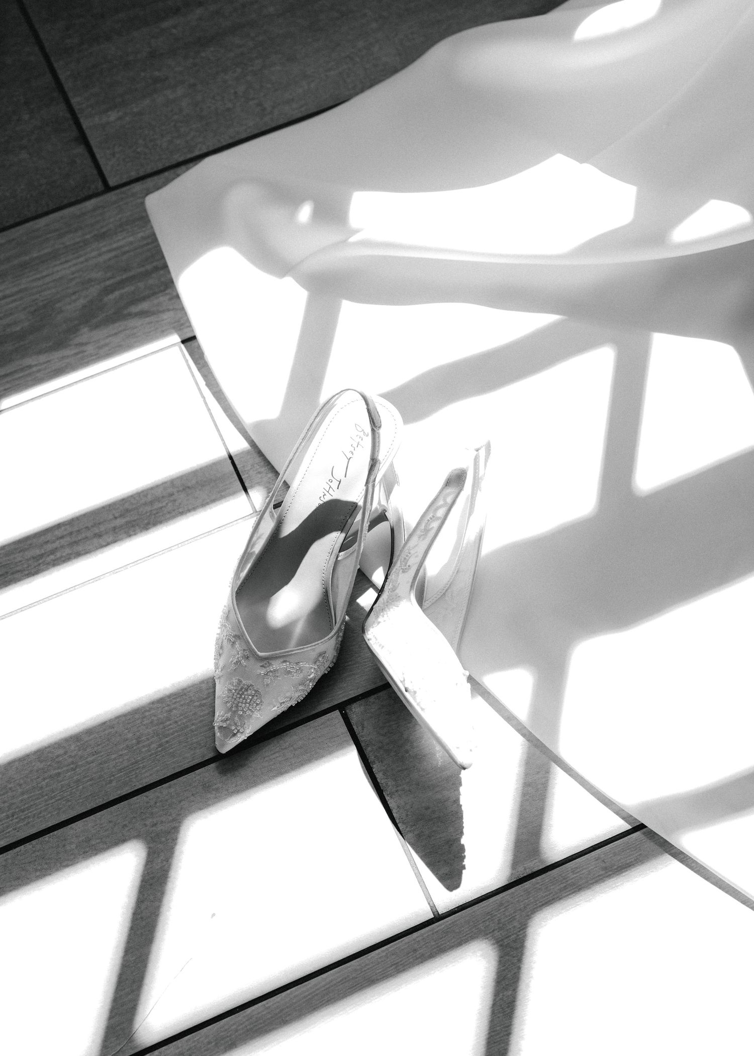 Black and white photo of elegant lace bridal heels on a wooden floor with dramatic light and shadow patterns.