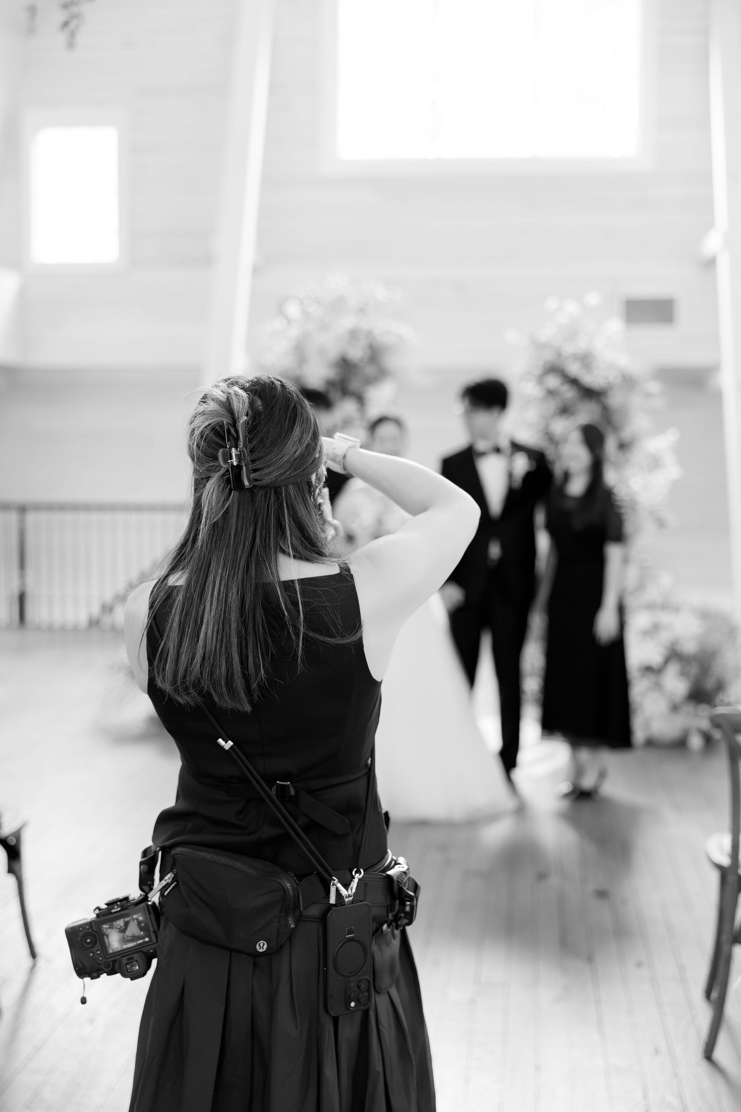Black and white photo of a female wedding photographer capturing a bride and groom posing under a floral arch.