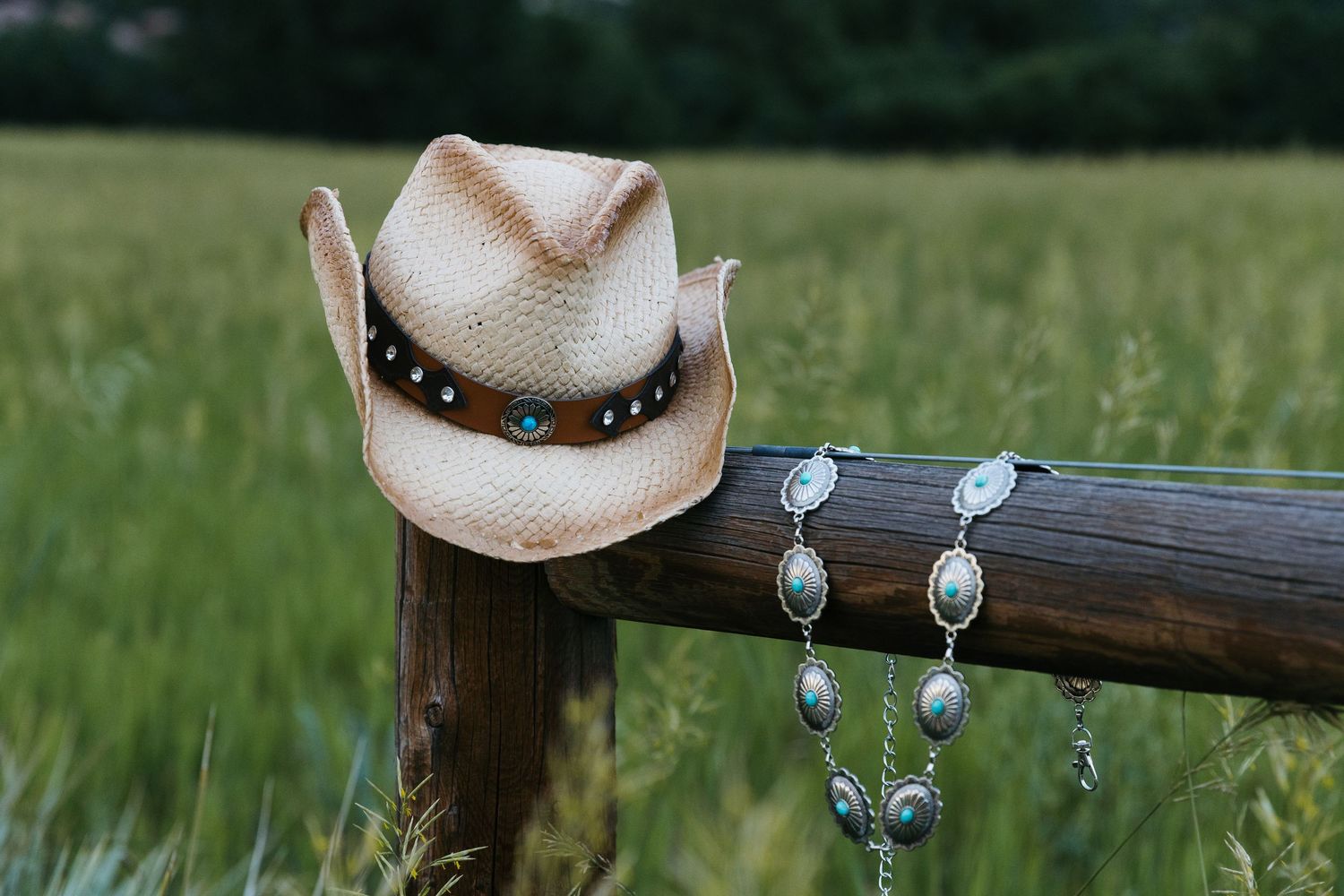 Straw cowboy hat with turquoise studded band and silver concho necklace draped on rustic wooden fence post in green field.