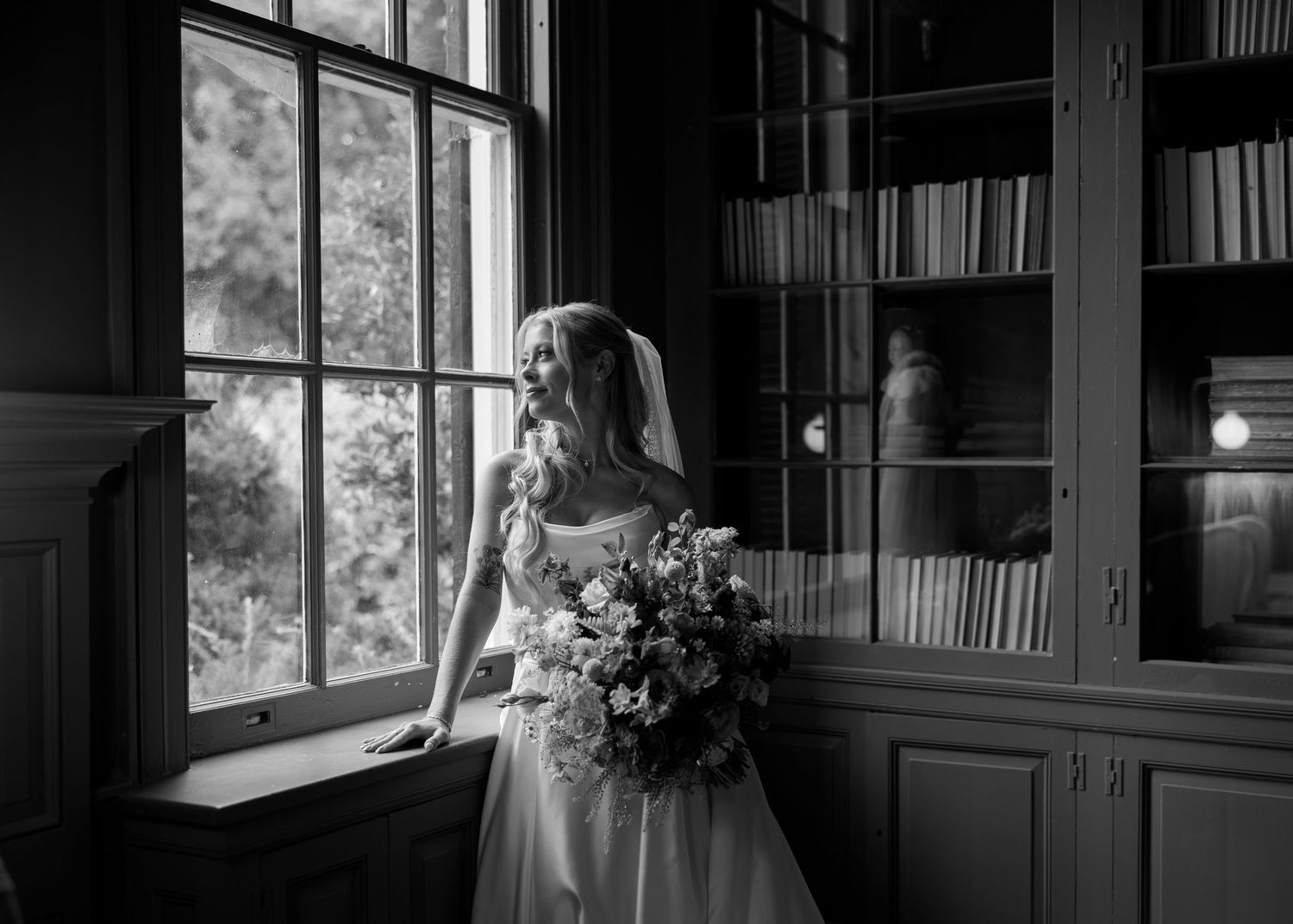 Bride in wedding dress holding bouquet stands by large window in elegant dark library, black and white photo.