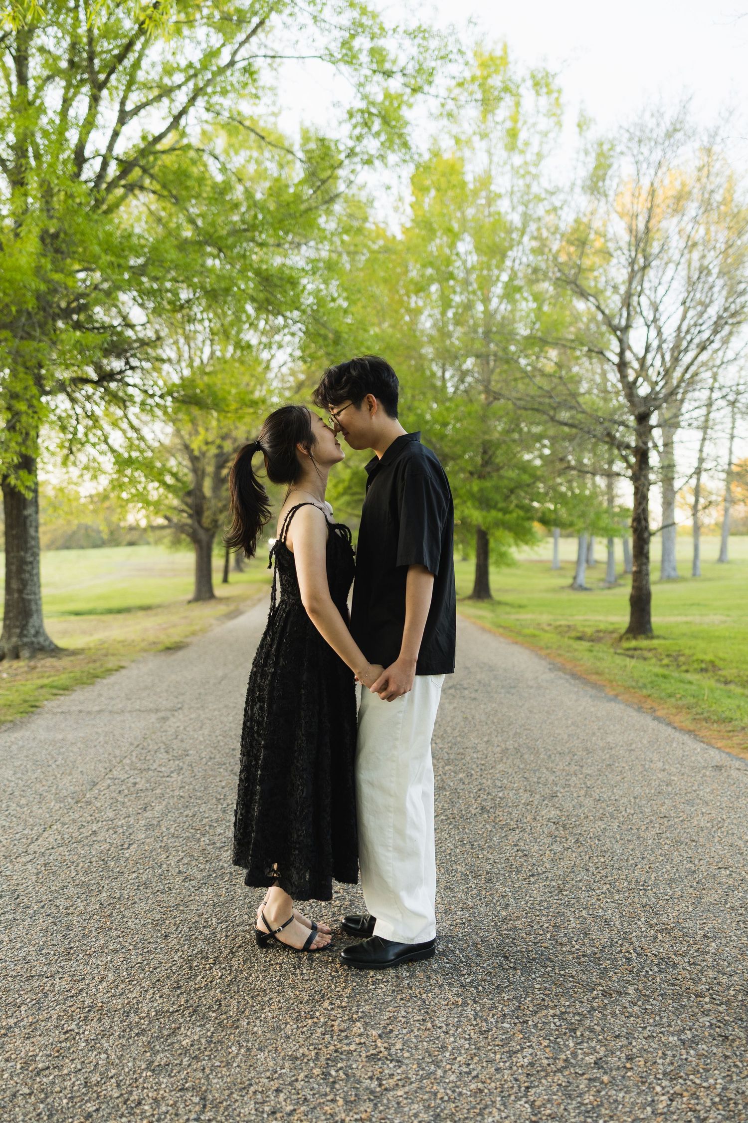 A couple stands close together holding hands on a park path, sharing an intimate moment surrounded by green trees.