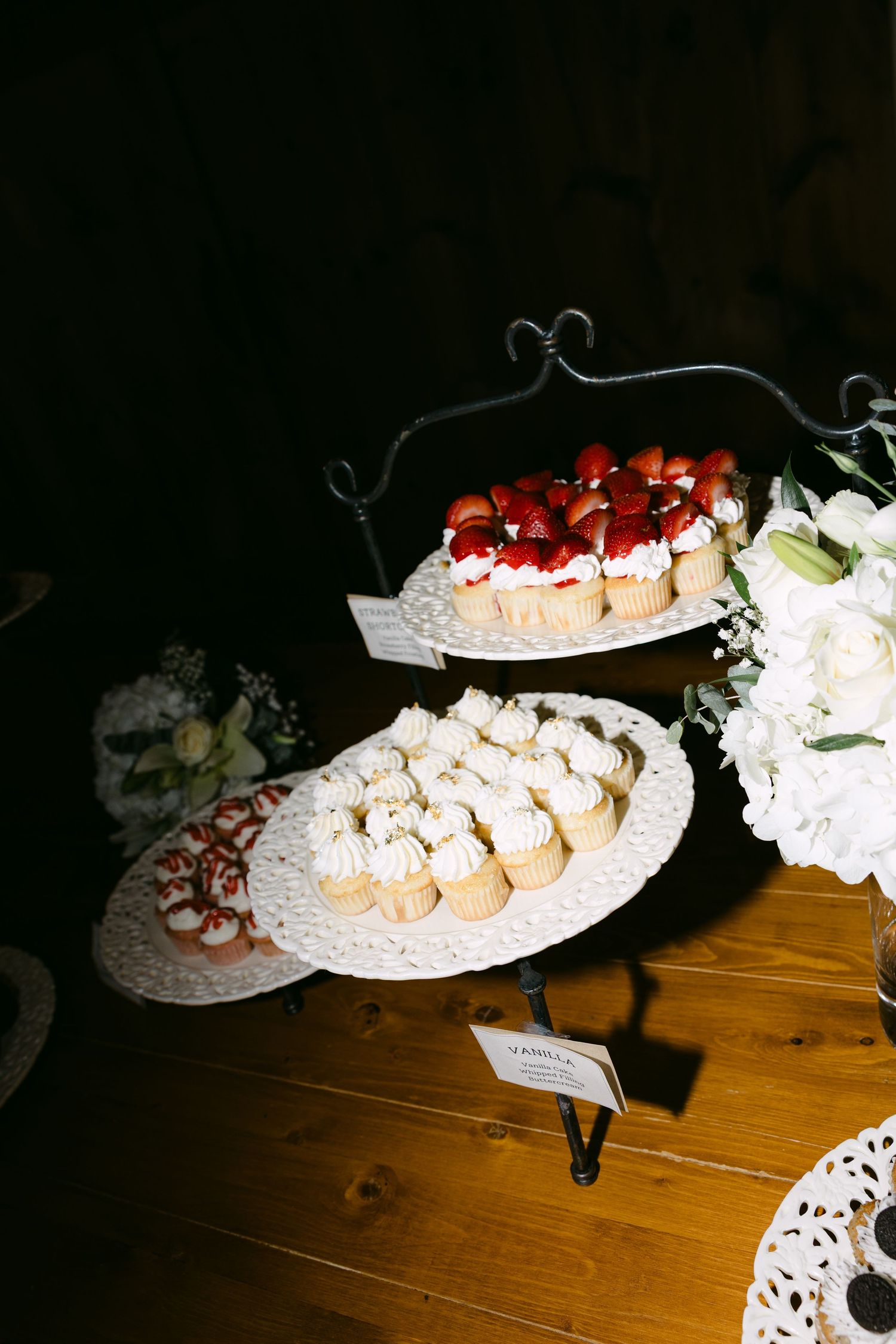 Two-tiered dessert stand with vanilla cupcakes and strawberry-topped treats beside white hydrangeas at a wedding reception.
