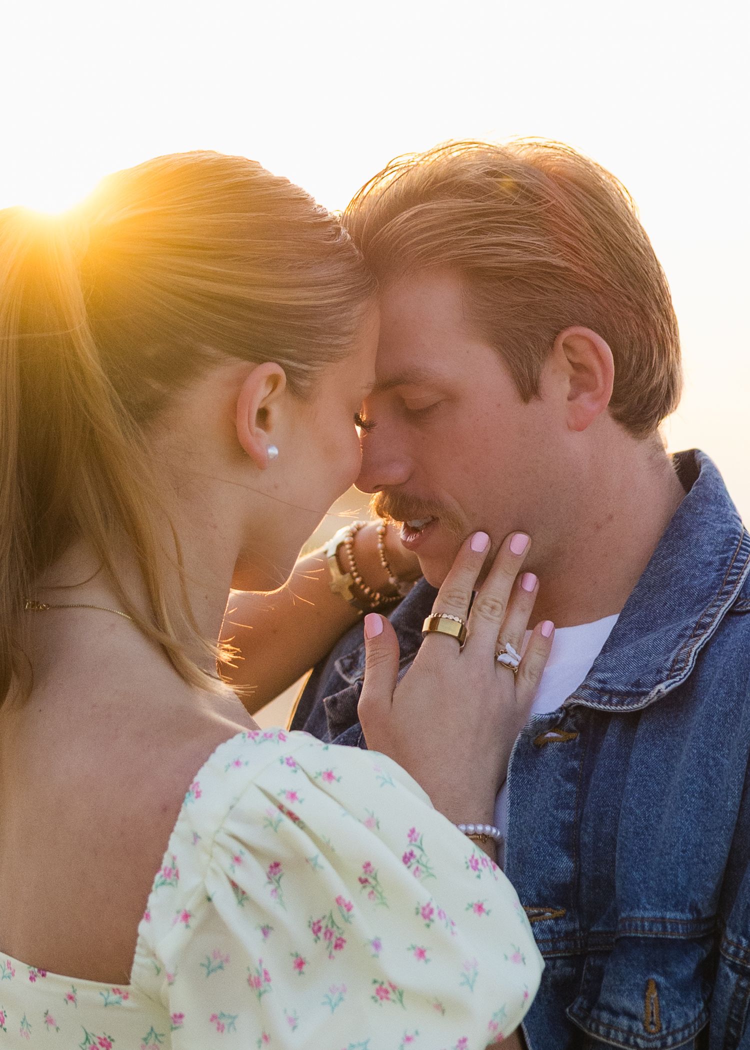 Couple sharing an intimate moment at golden sunset, woman touching mans face with engagement ring visible.
