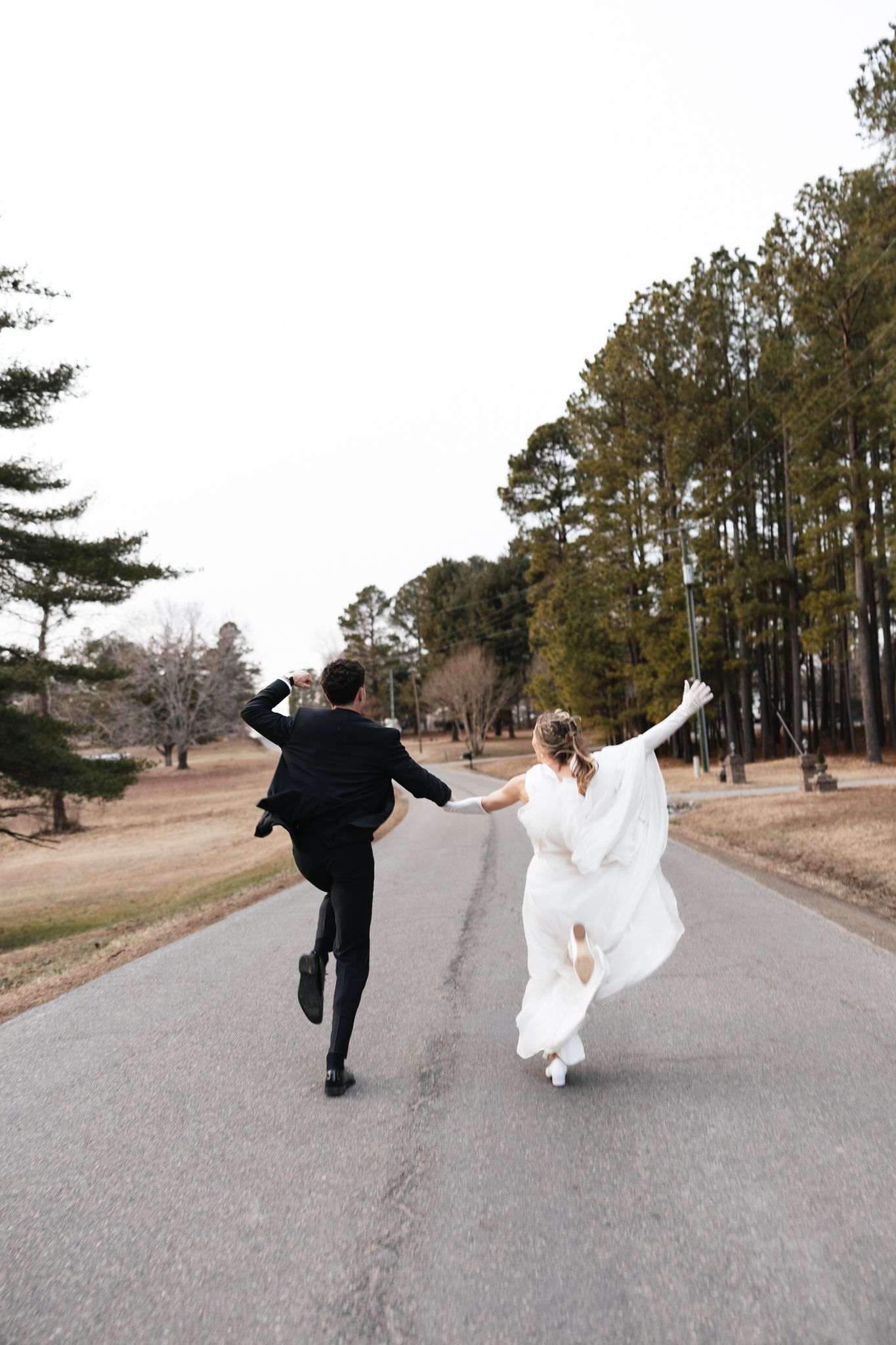 Bride and groom joyfully skip down a tree-lined road holding hands during outdoor wedding portraits.