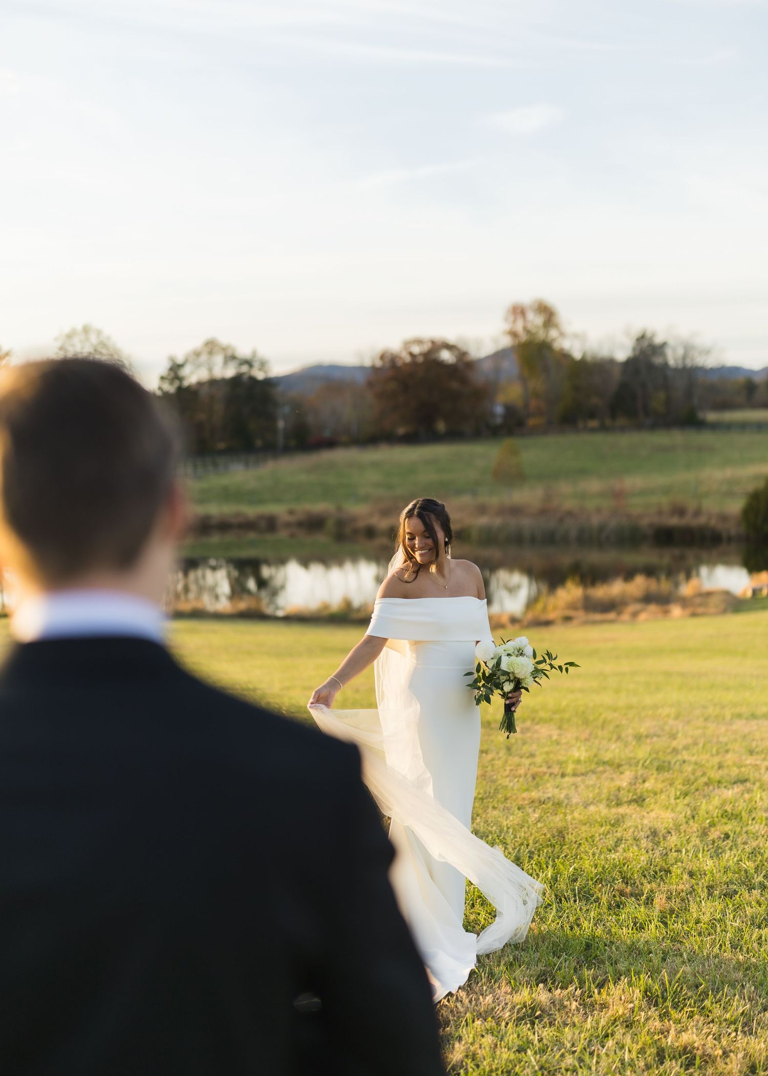 Radiant bride in off-shoulder gown walks toward groom across a sunlit field with a pond and mountains behind her.