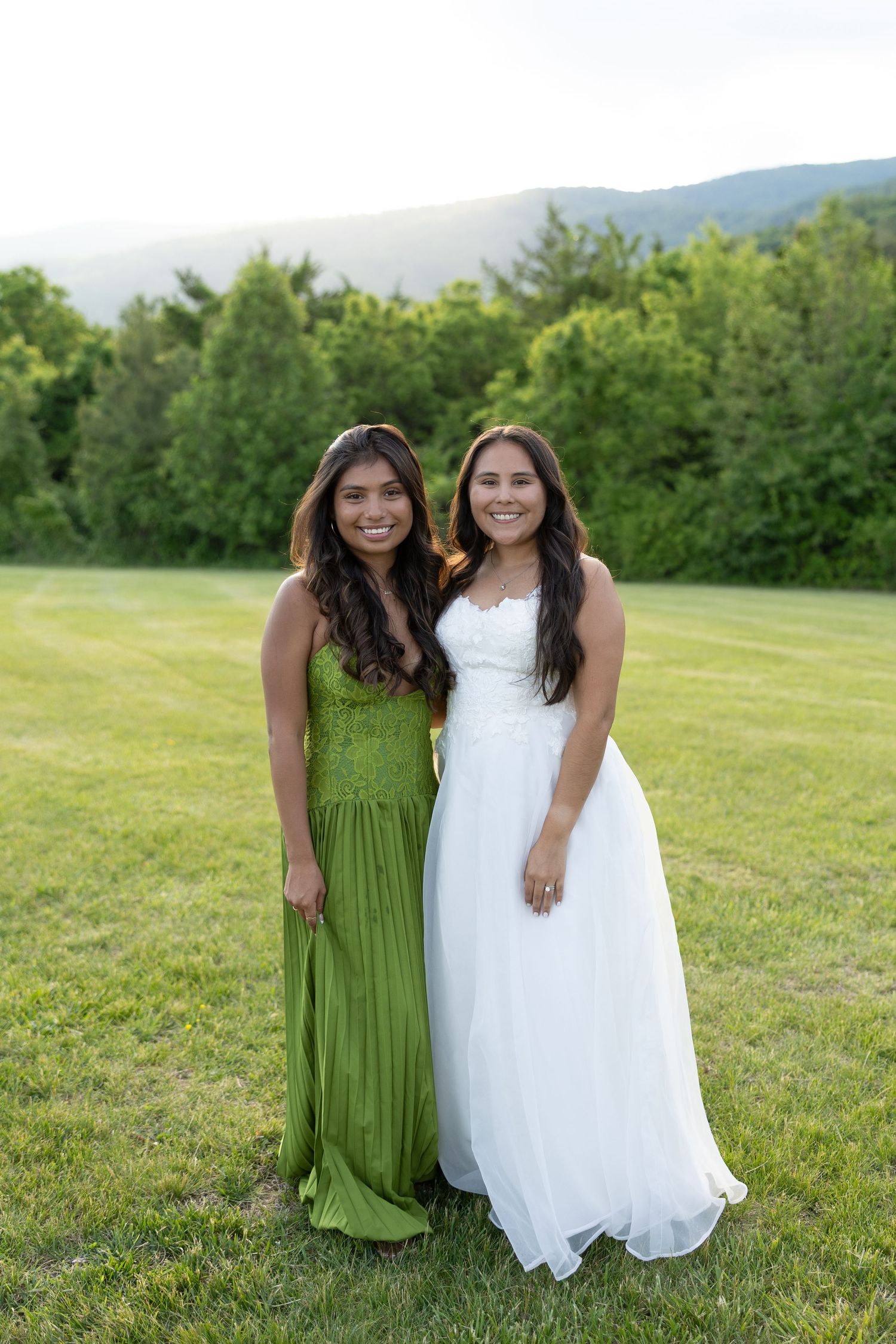 Two young women in formal gowns, one green and one white, posing outdoors on green grass with trees and mountains.