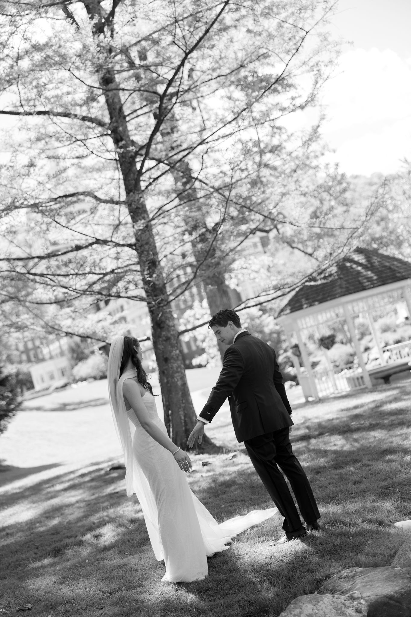 Black and white photo of bride and groom holding hands walking under trees near a gazebo.