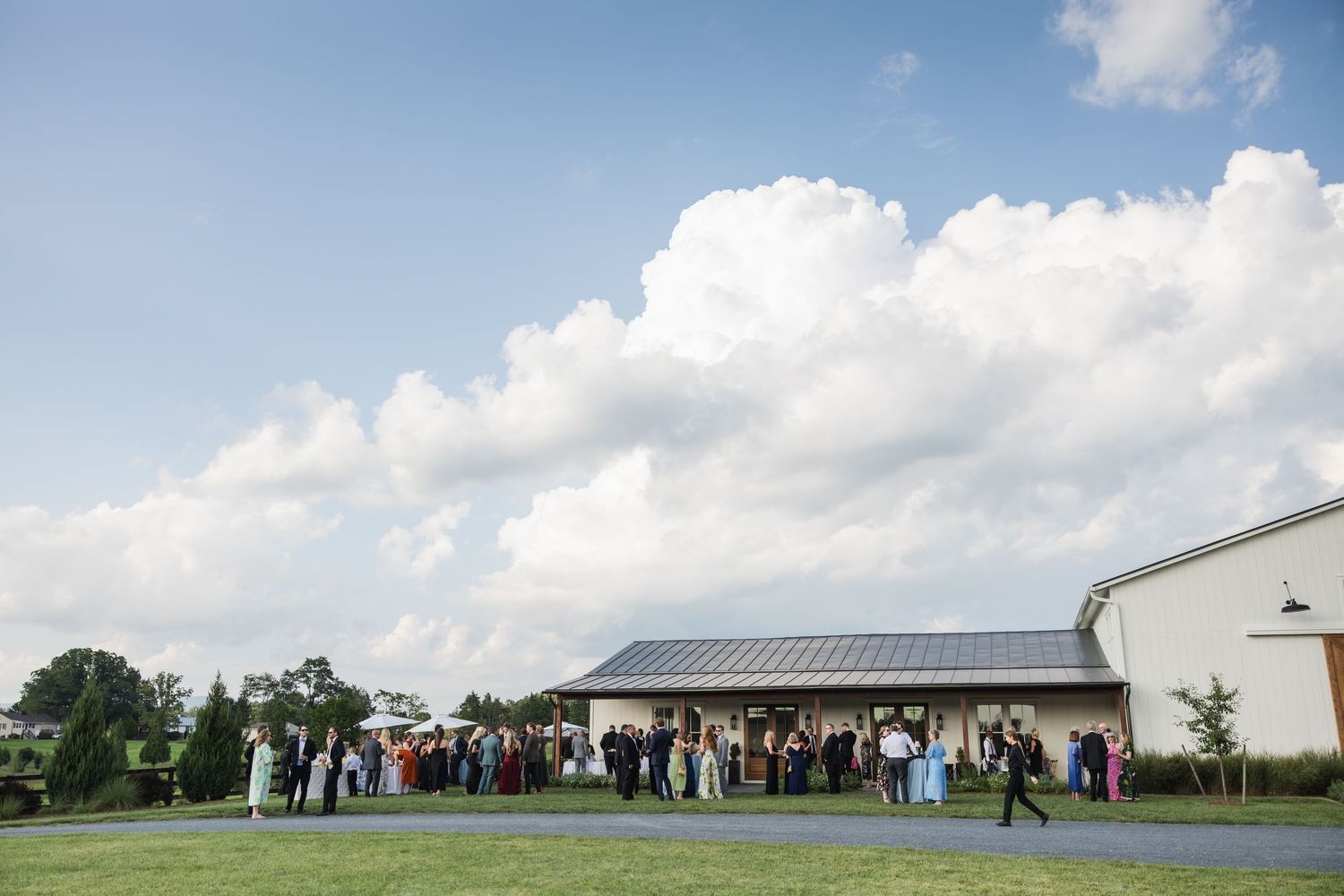 Outdoor wedding reception with guests gathered on green lawn beside modern barn venue under dramatic cloudy blue sky.