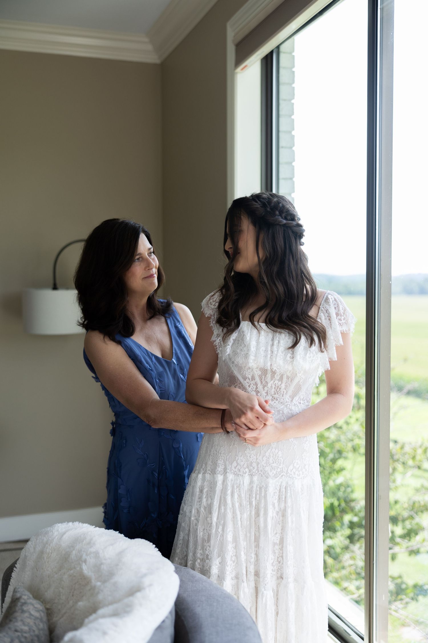 Bride in lace wedding dress sharing a tender moment with her mother by a bright window.