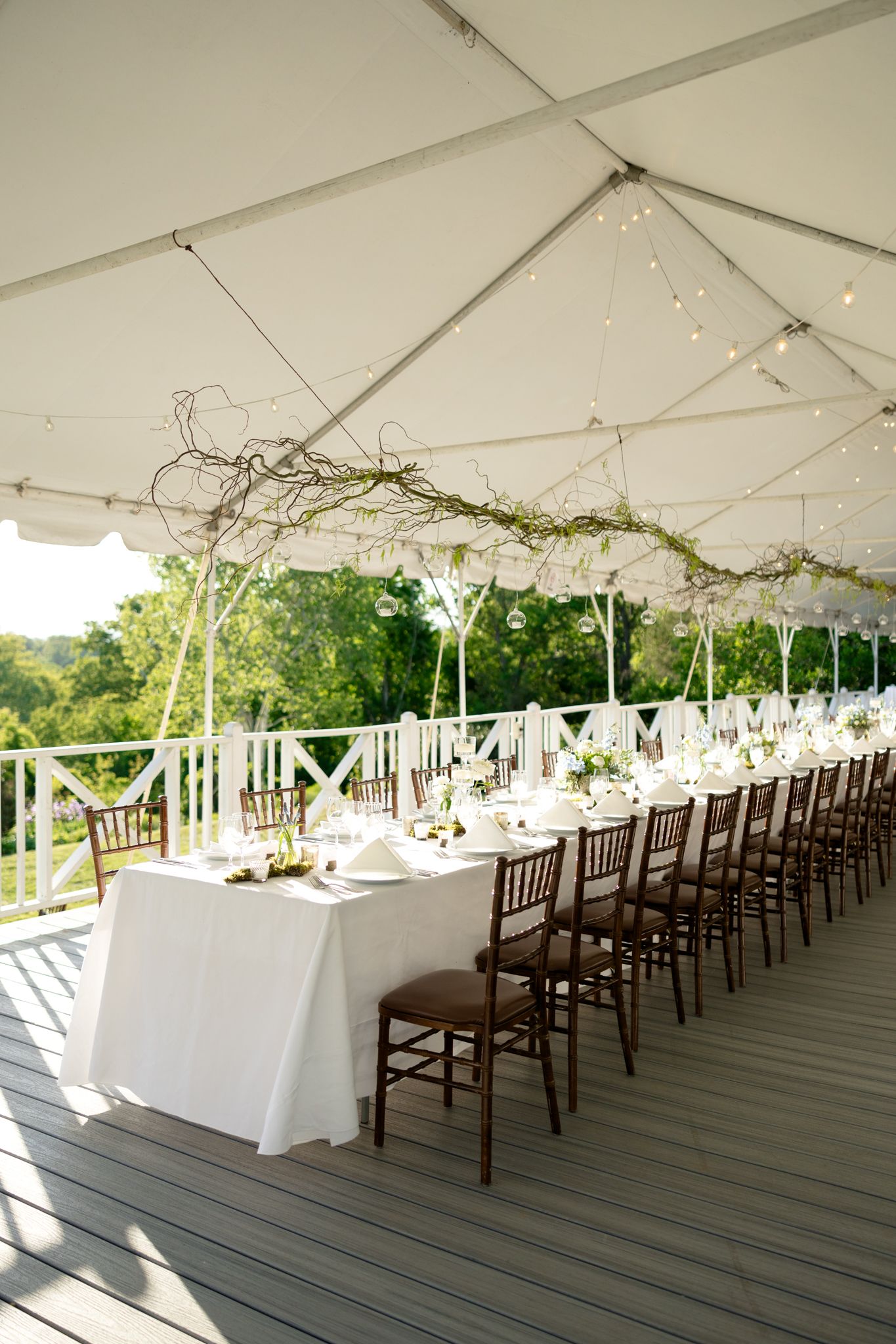 Elegant outdoor wedding reception under white tent with long table, brown chiavari chairs, and floral branch centerpieces.