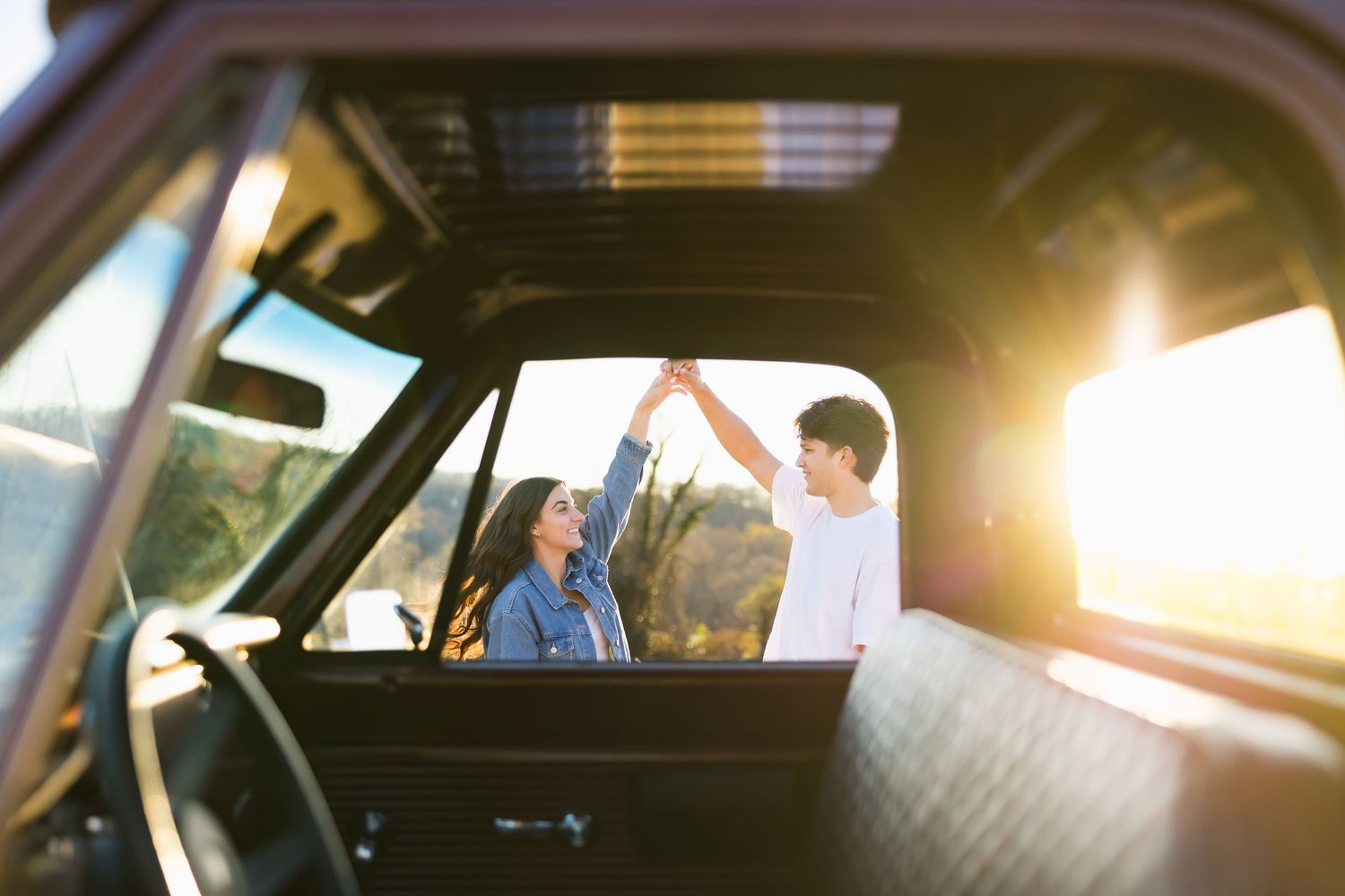 Joyful couple holding hands and dancing beside a classic truck with bright golden sunset light streaming through.