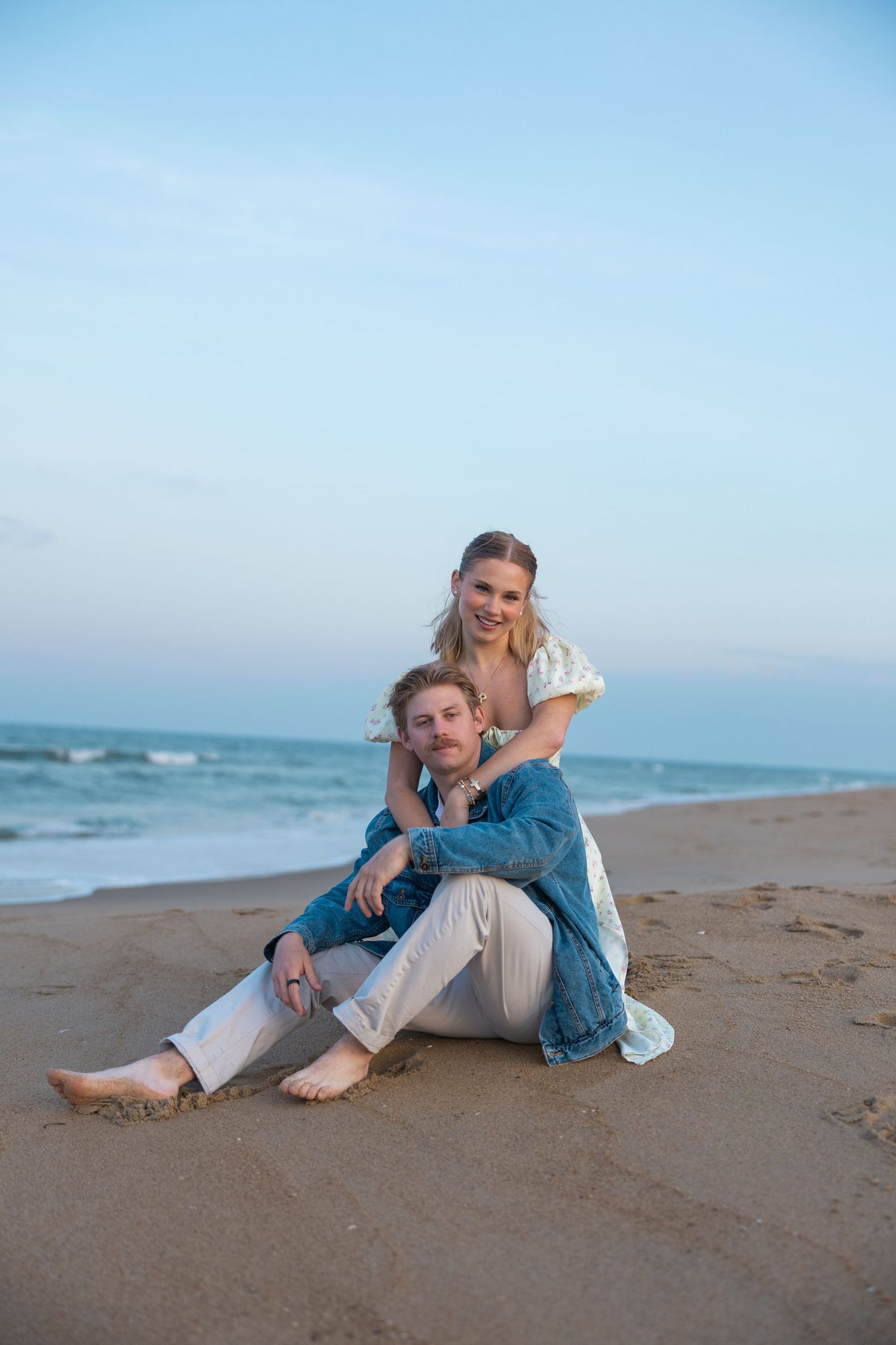 Happy couple sitting together on sandy beach at dusk, woman embracing man from behind.