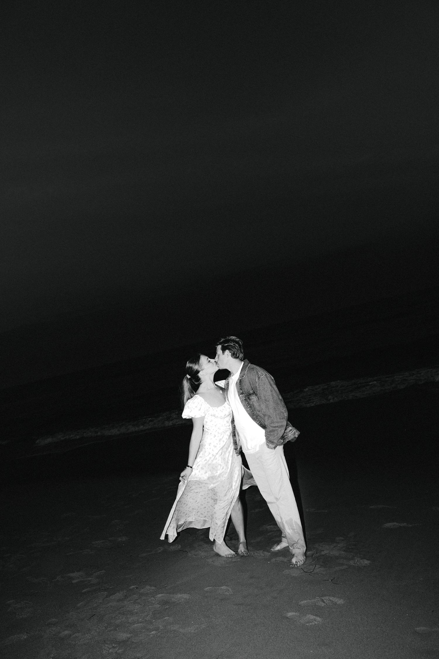 Couple shares a kiss on dark beach at night in romantic black and white photo.